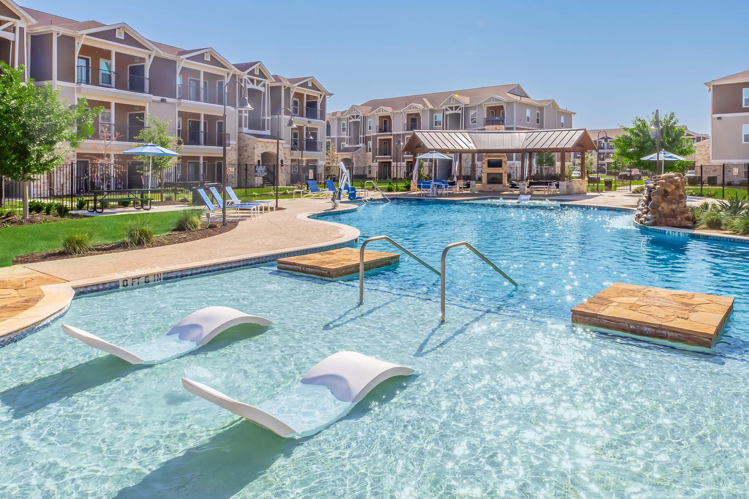 A bright outdoor swimming pool surrounded by lounge chairs and umbrellas, with modern apartment buildings in the background. The pool features a shallow section with two white lounge chairs partially submerged in the water, inviting relaxation on a sunny day. Lush green landscaping enhances the inviting atmosphere.