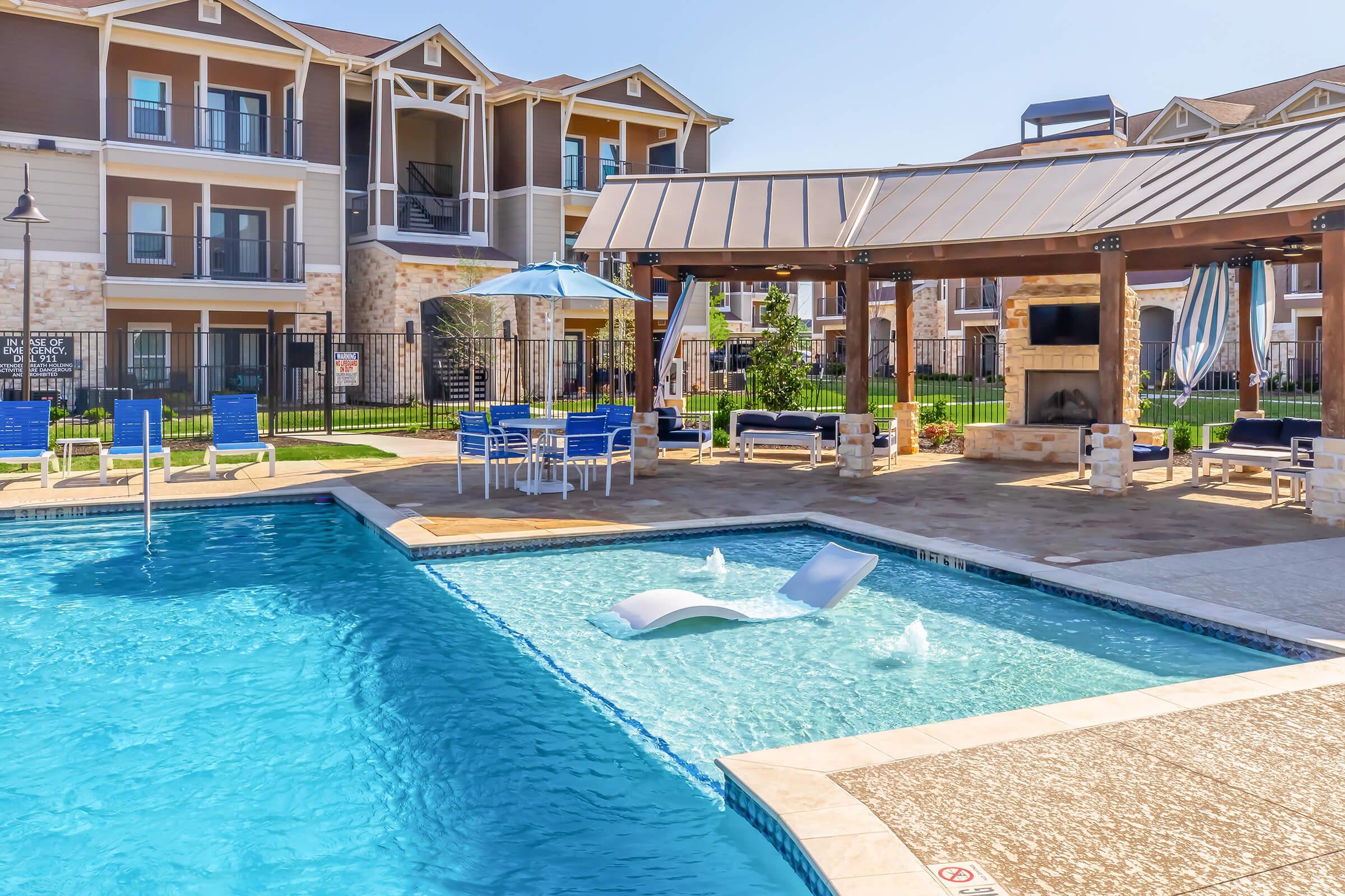 A bright, inviting swimming pool area featuring a clear blue pool with lounge chairs and a reclining float. Nearby, a shaded gazebo with seating and an outdoor fireplace overlooks the pool. Lush green grass and well-maintained landscaping surround the space, creating a relaxing atmosphere in an apartment complex.