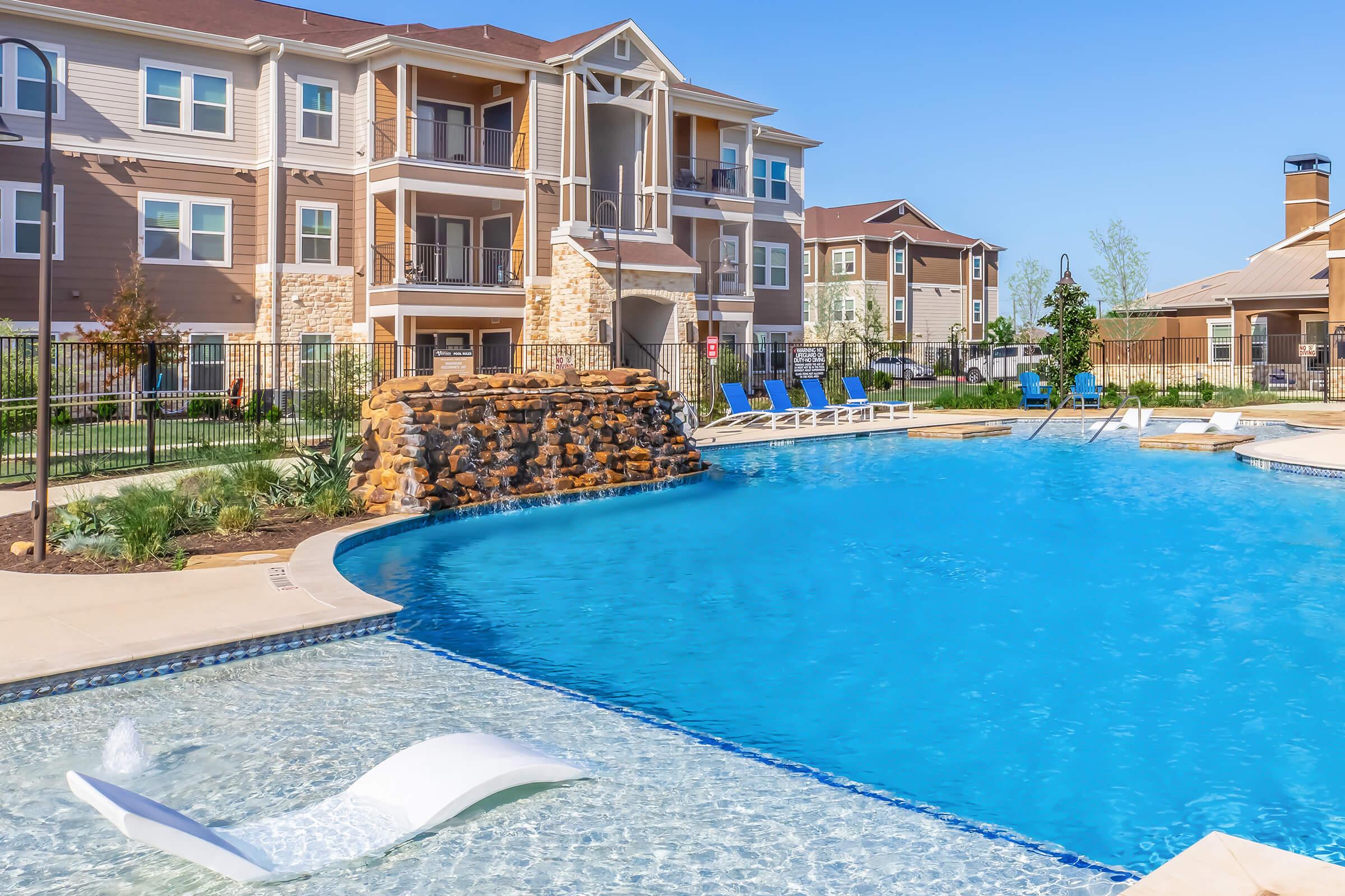 A large outdoor swimming pool with a rock formation waterfall, surrounded by lounge chairs and modern apartment buildings in the background. The area is well-maintained with green landscaping under a clear blue sky. The pool features both shallow and deep sections.