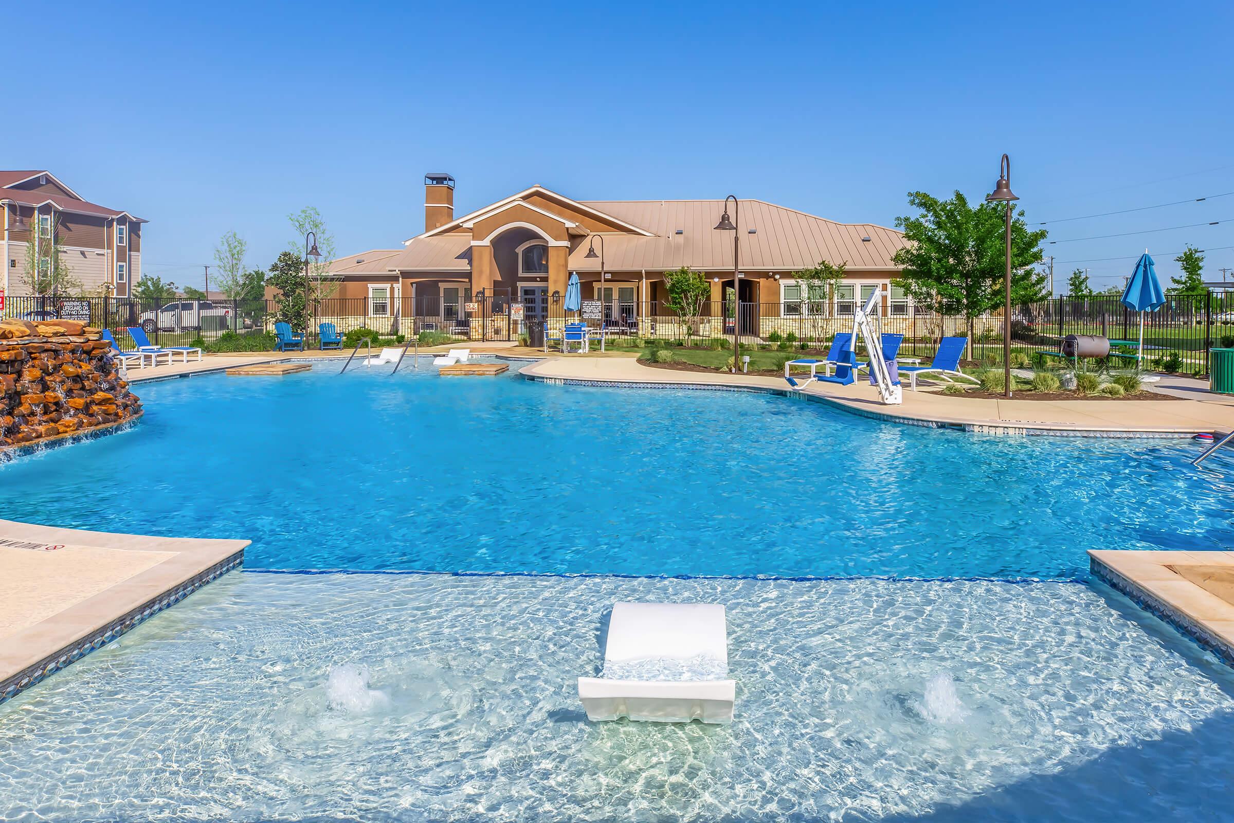 A spacious outdoor swimming pool with clear blue water, surrounded by sun loungers and umbrellas. In the background, there is a modern building with a brown roof and a stone feature. Lush green trees and a well-maintained lawn enhance the inviting atmosphere. Clear blue sky above completes the scene.