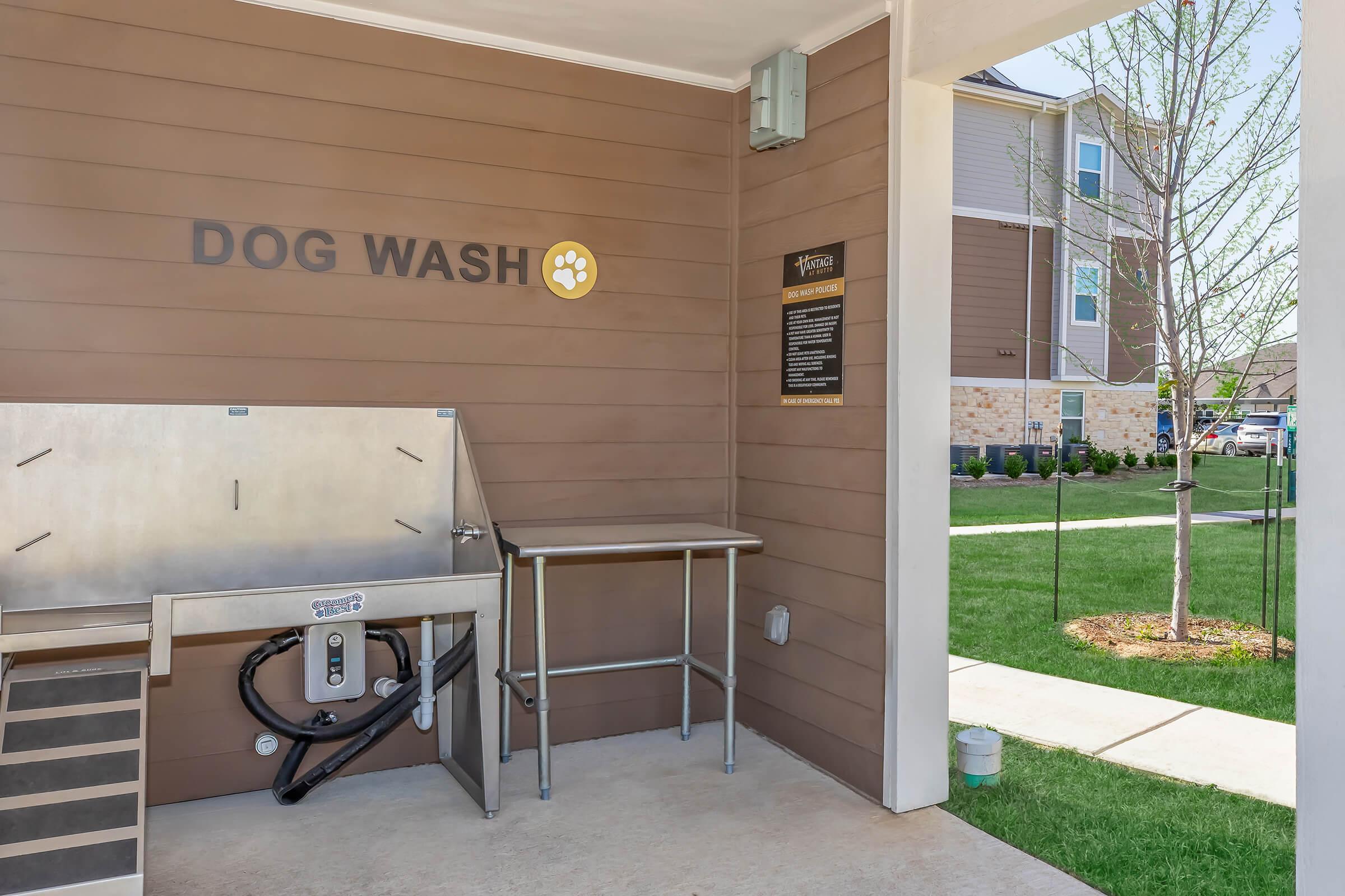 Outdoor dog wash station with a stainless steel tub, attached hose, and nearby grooming supplies. The wall features "DOG WASH" text and a small sign with instructions. Surrounding area has green grass and a glimpse of residential buildings in the background.