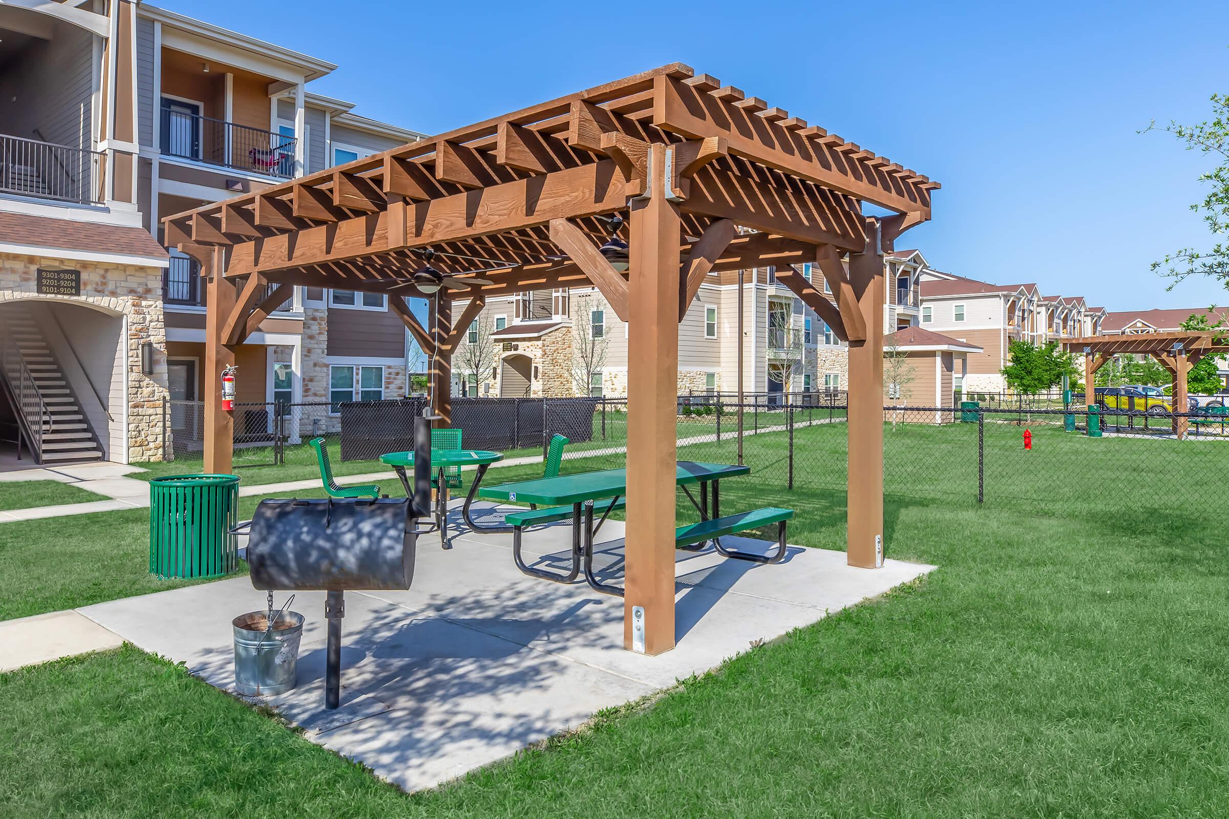 A wooden pergola with picnic tables and a grill is situated in a grassy area, surrounded by an apartment complex. The clear blue sky provides a bright backdrop to the setting. Trash can visible nearby, and a fence delineates the space. Overall, a welcoming outdoor recreational area.
