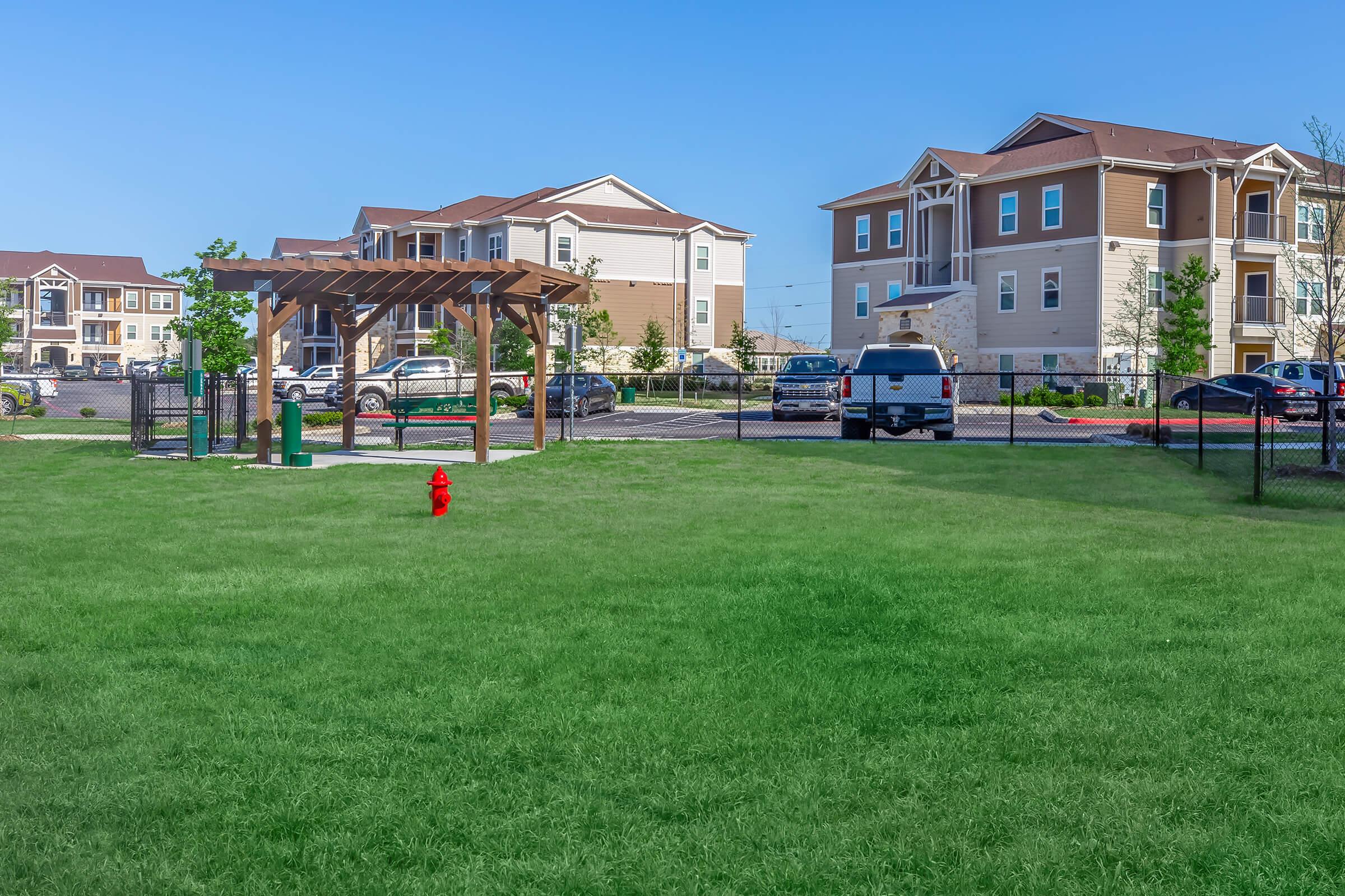A grassy area featuring a red fire hydrant and a wooden gazebo. In the background, there are several residential buildings and parked cars. The sky is clear and blue, creating a bright and inviting atmosphere. The area is enclosed with a fence, suggesting it is part of a community space.