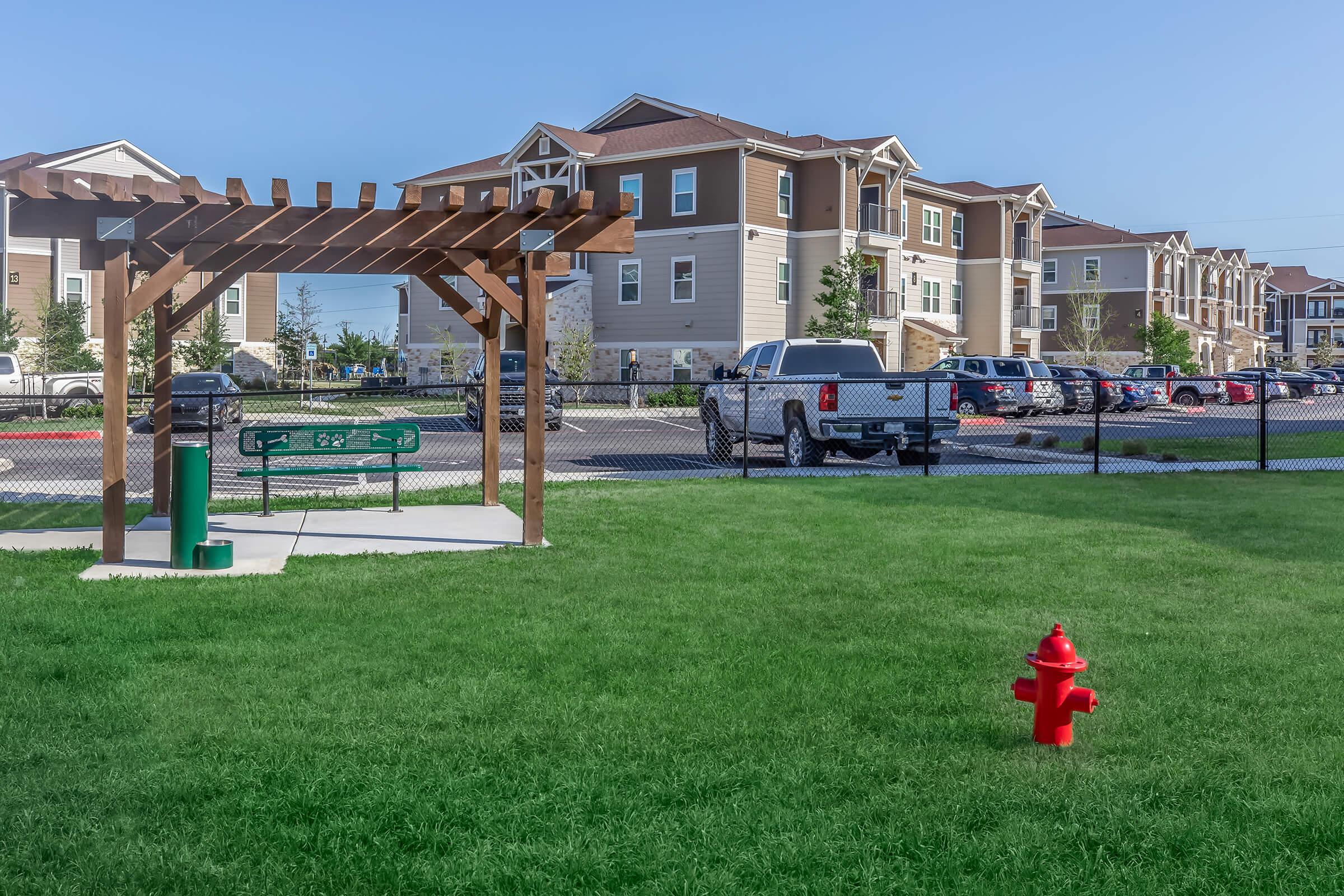 A grassy area in a residential community featuring a wooden pergola with benches, a green garbage can, and a red fire hydrant. In the background, modern apartment buildings and parked cars are visible, under a clear blue sky. The setting suggests a pet-friendly environment.