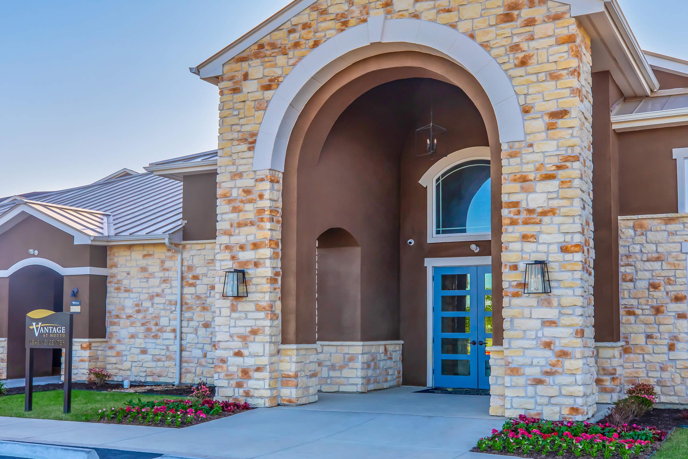 Entrance of a modern building featuring a large archway and a blue door. The façade combines light-colored stone and brown elements. Flanking the entrance are two lamps on either side and landscaped flower beds with colorful plants. A sign reads "Vantage."