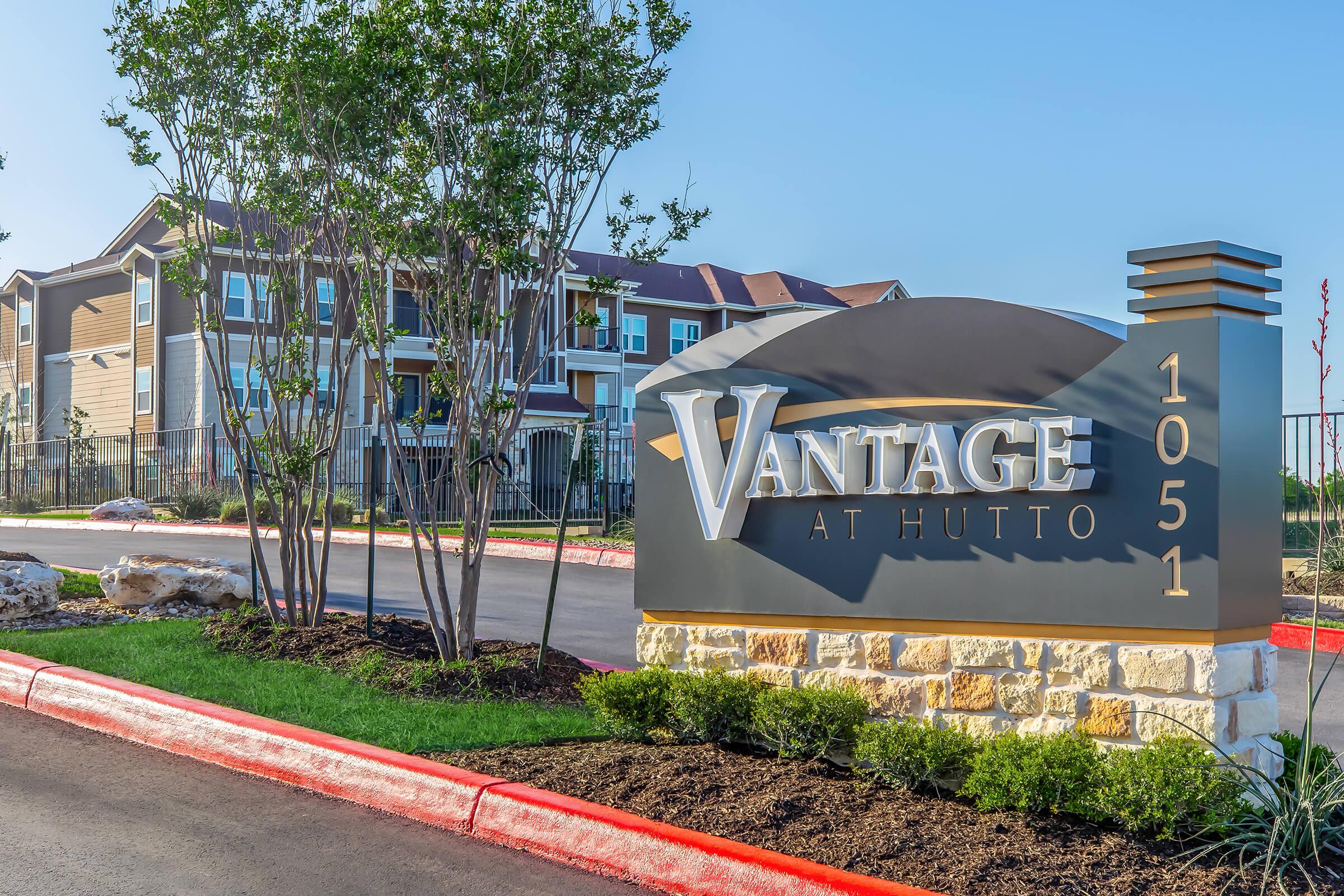 Sign for Vantage at Hutto, a residential apartment community, featuring the name prominently displayed. The sign is situated in a landscaped area with trees and shrubs. In the background, modern apartment buildings are visible against a clear blue sky. The scene conveys a welcoming atmosphere for potential residents.