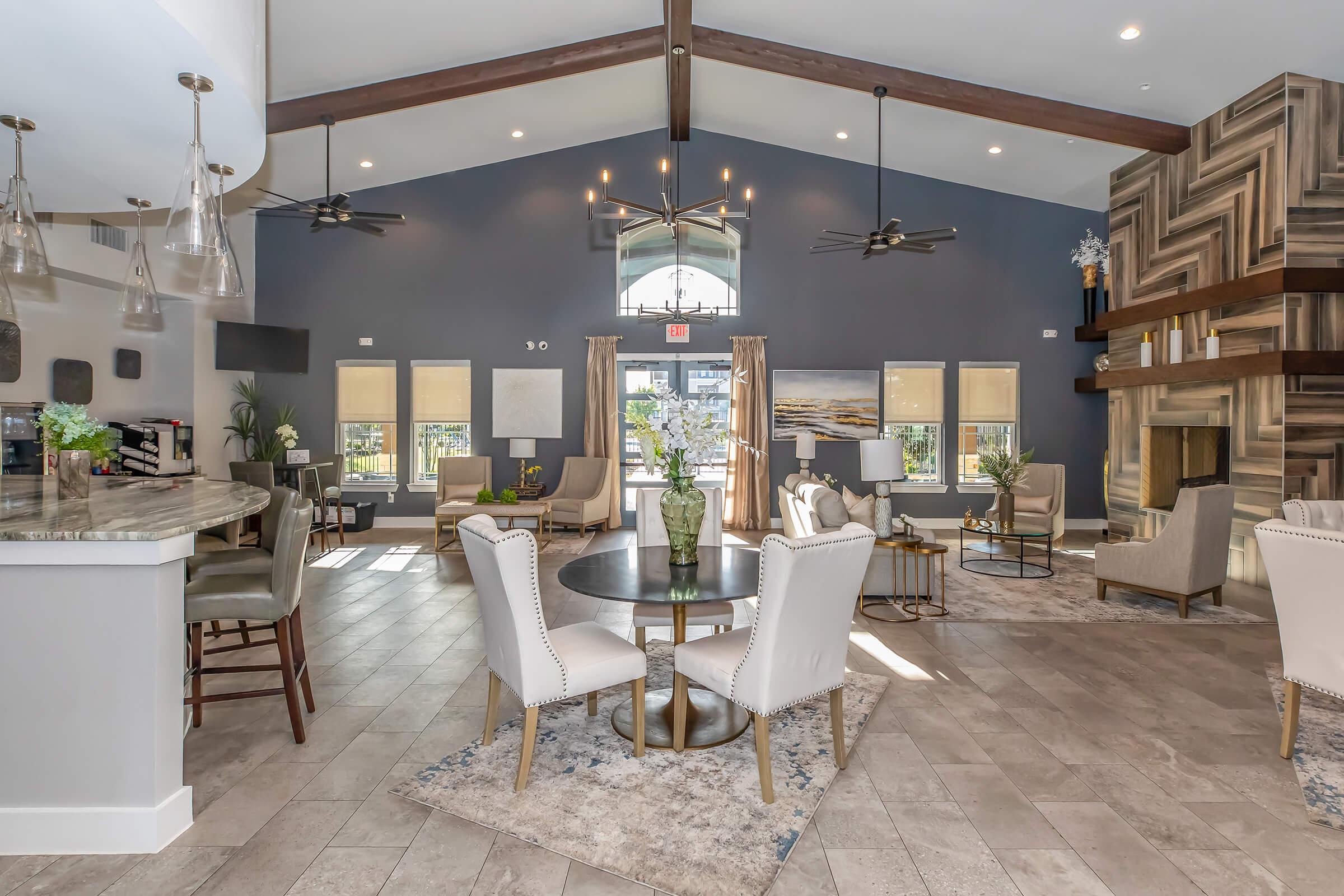 A spacious, modern lounge area featuring high ceilings, large windows, and elegant furniture. The space includes a cozy seating arrangement with armchairs and a round table, a stylish bar counter, and an eye-catching stone fireplace. Decorative elements and plants enhance the inviting atmosphere.