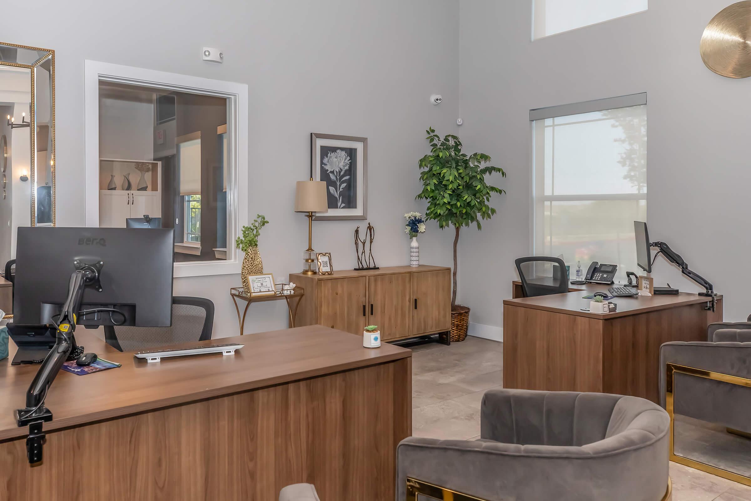 A modern office space featuring two wooden desks with computers, stylish chairs, and contemporary decor. There is a large window allowing natural light, a potted plant, and decorative artwork on the walls. The design is sleek and inviting, with a neutral color palette.