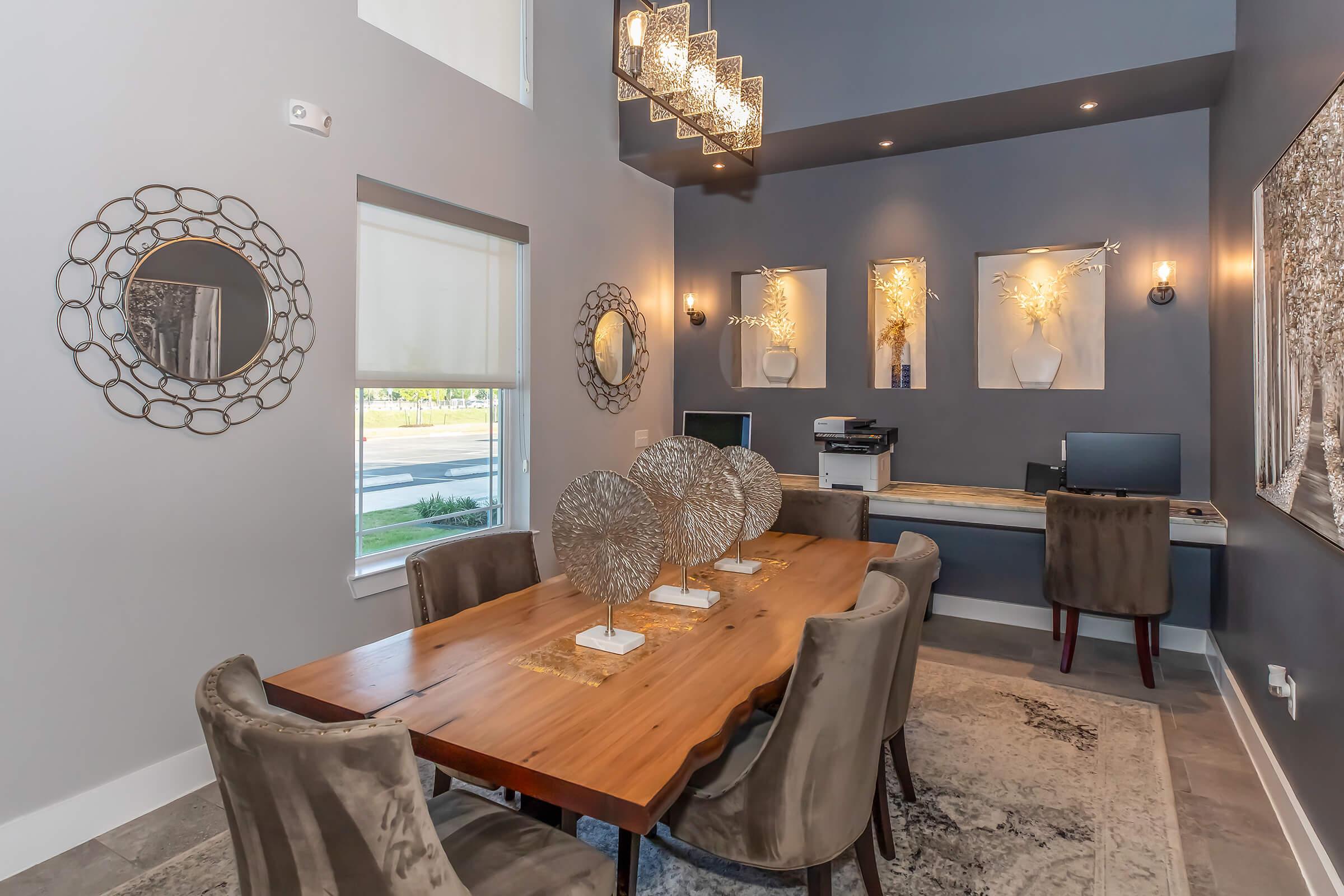 A modern dining area with a wooden table surrounded by plush chairs. The walls are painted gray and feature decorative mirrors. A stylish chandelier hangs above, and there are decorative vases on the table. A computer station is visible in the corner of the room, enhancing the contemporary design.