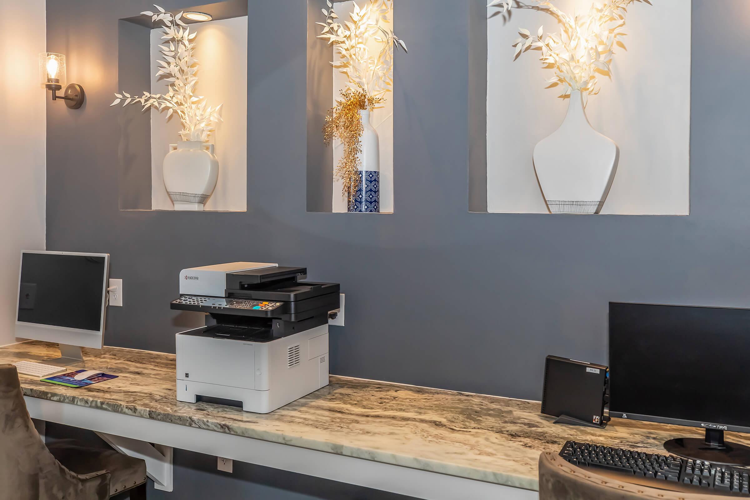 A modern office space featuring a dark blue wall with decorative vases and greenery. In the foreground, there's a large marble-top desk with a printer, computer monitors, and a keyboard. The atmosphere is sleek and contemporary, ideal for a productive environment.