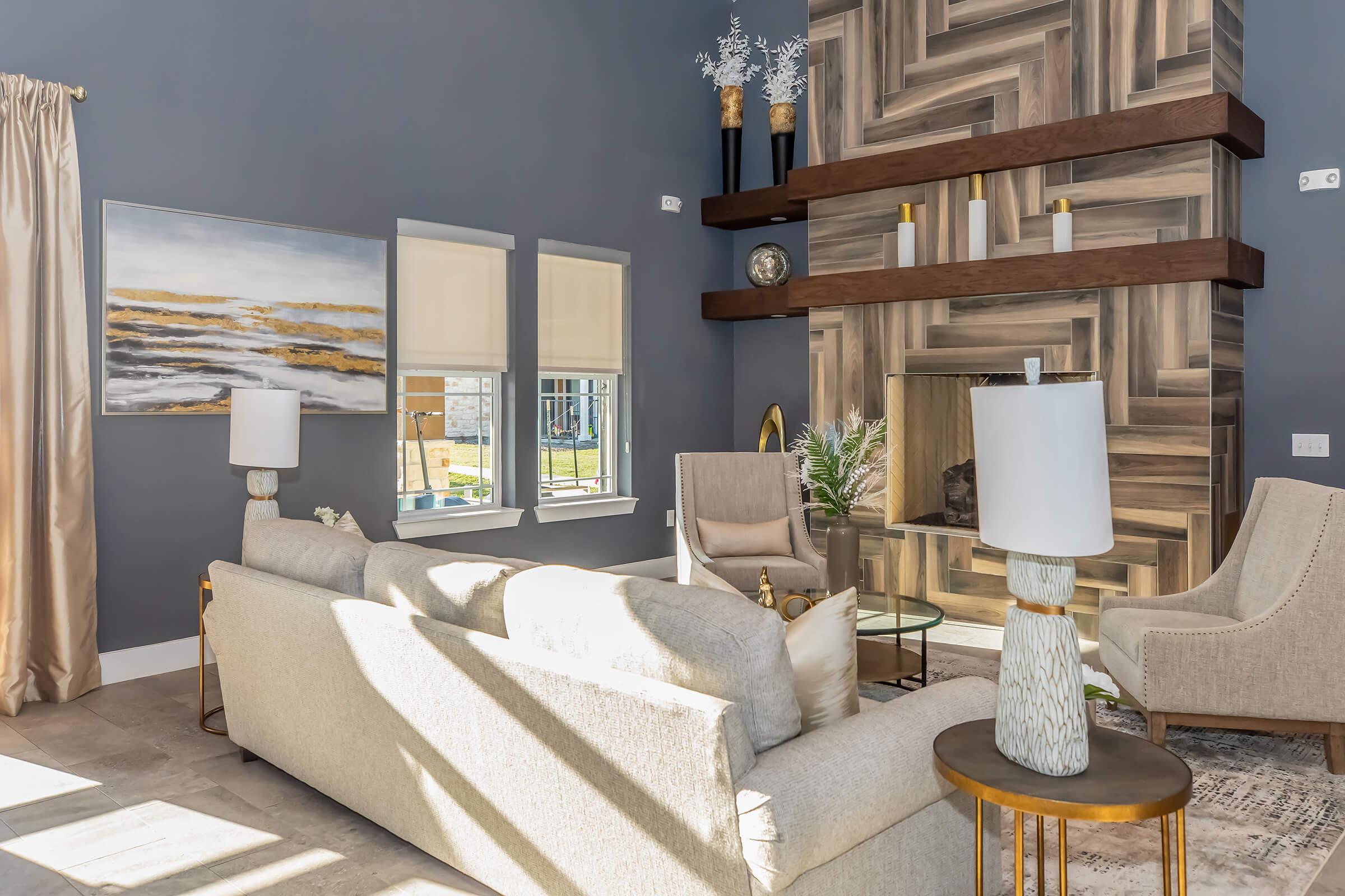 A modern living room featuring a light-colored sofa and armchairs, a wooden accent wall with a built-in fireplace, and decorative items. Large windows allow natural light to illuminate the space, while a stylish painting adds a touch of color. Cozy and elegant atmosphere with warm accents.