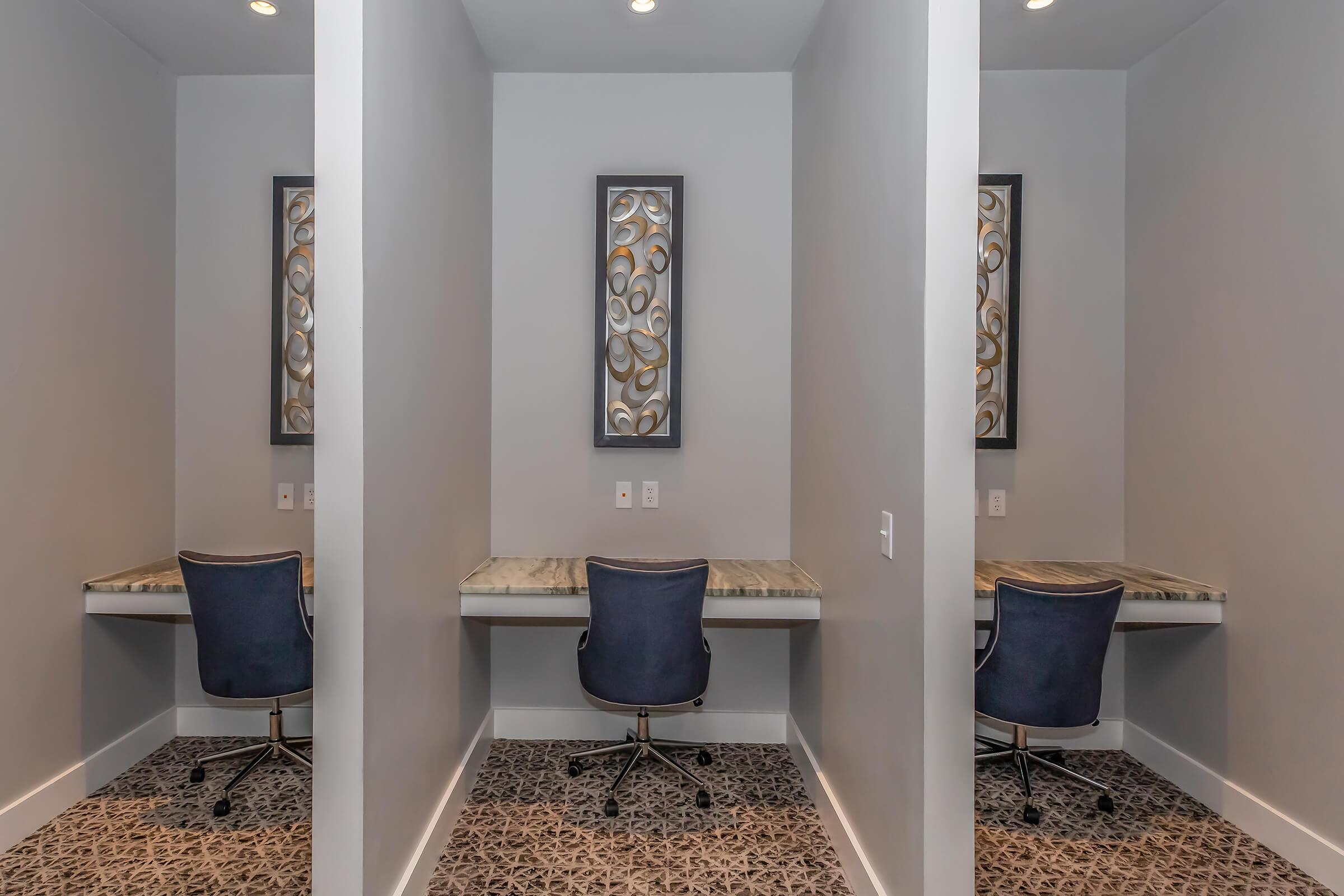 A modern workspace featuring two individual desks with blue rolling chairs, separated by light gray partitions. Each desk has a minimalist design, and there are decorative framed pieces on the walls. The flooring has a patterned design, enhancing the contemporary aesthetic of the area.