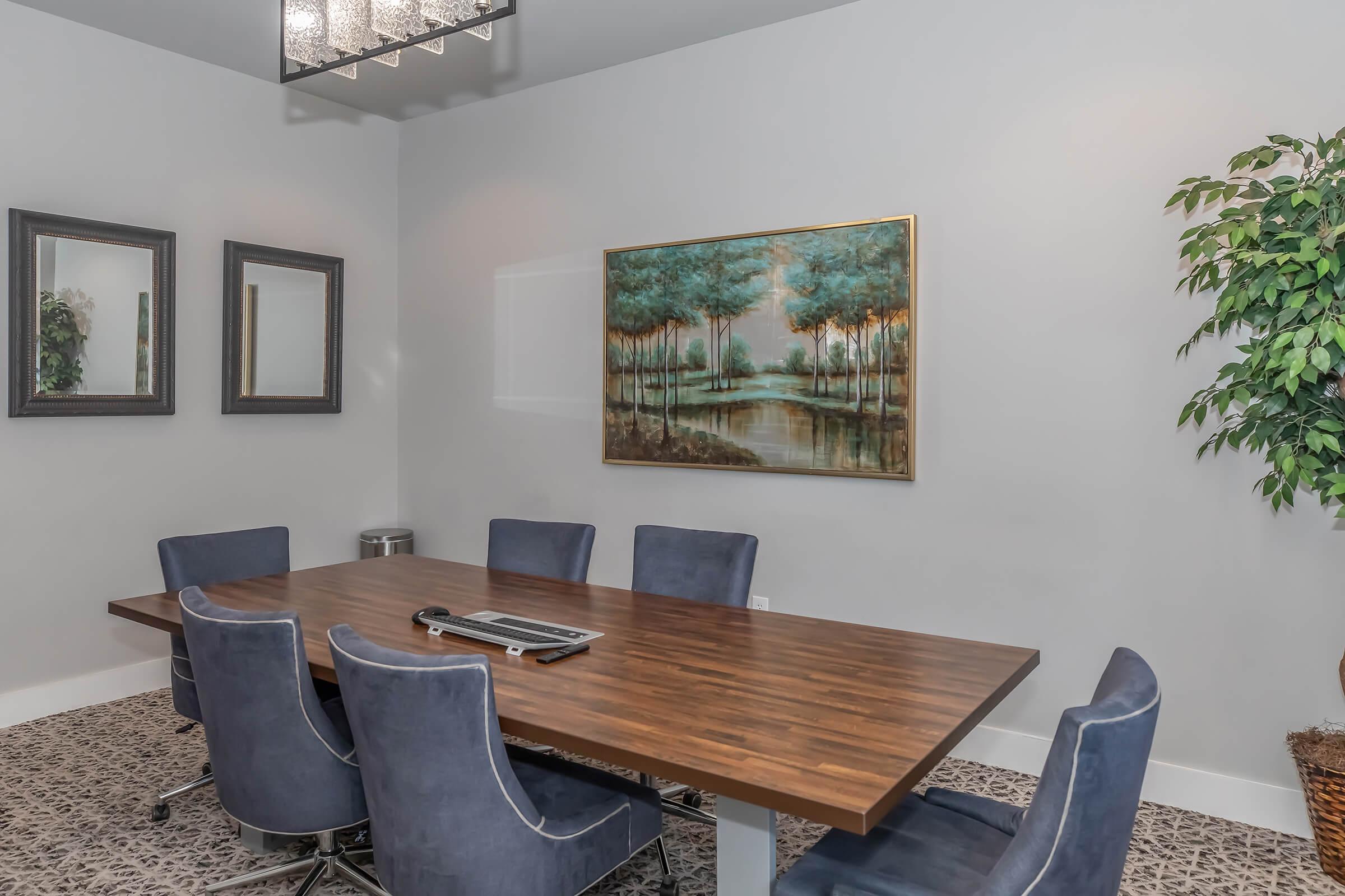 A modern conference room featuring a large wooden table surrounded by six blue upholstered chairs. The room has light gray walls, a chandelier, a painting of trees by a lake, and two mirrors. A potted plant adds a touch of greenery to the space, creating a professional yet inviting atmosphere.