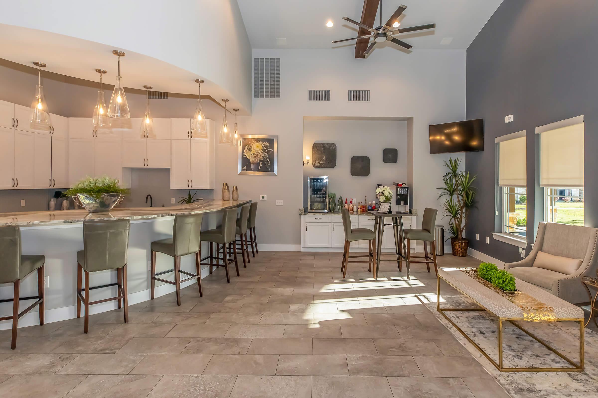 Modern open-concept living space featuring a kitchen bar with high stools, pendant lighting, and a stylish seating area. The room has large windows with natural light, chic decor, and a small table with plants, creating a welcoming atmosphere for relaxation and socializing.