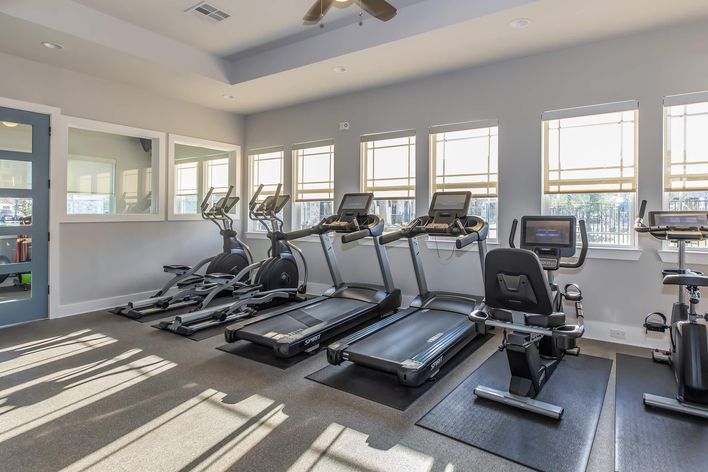 A modern gym interior featuring exercise equipment, including treadmills and elliptical machines, arranged against large windows. Natural light floods the space, creating a bright environment. The walls are painted in neutral colors, and the floor is covered with dark carpeting.