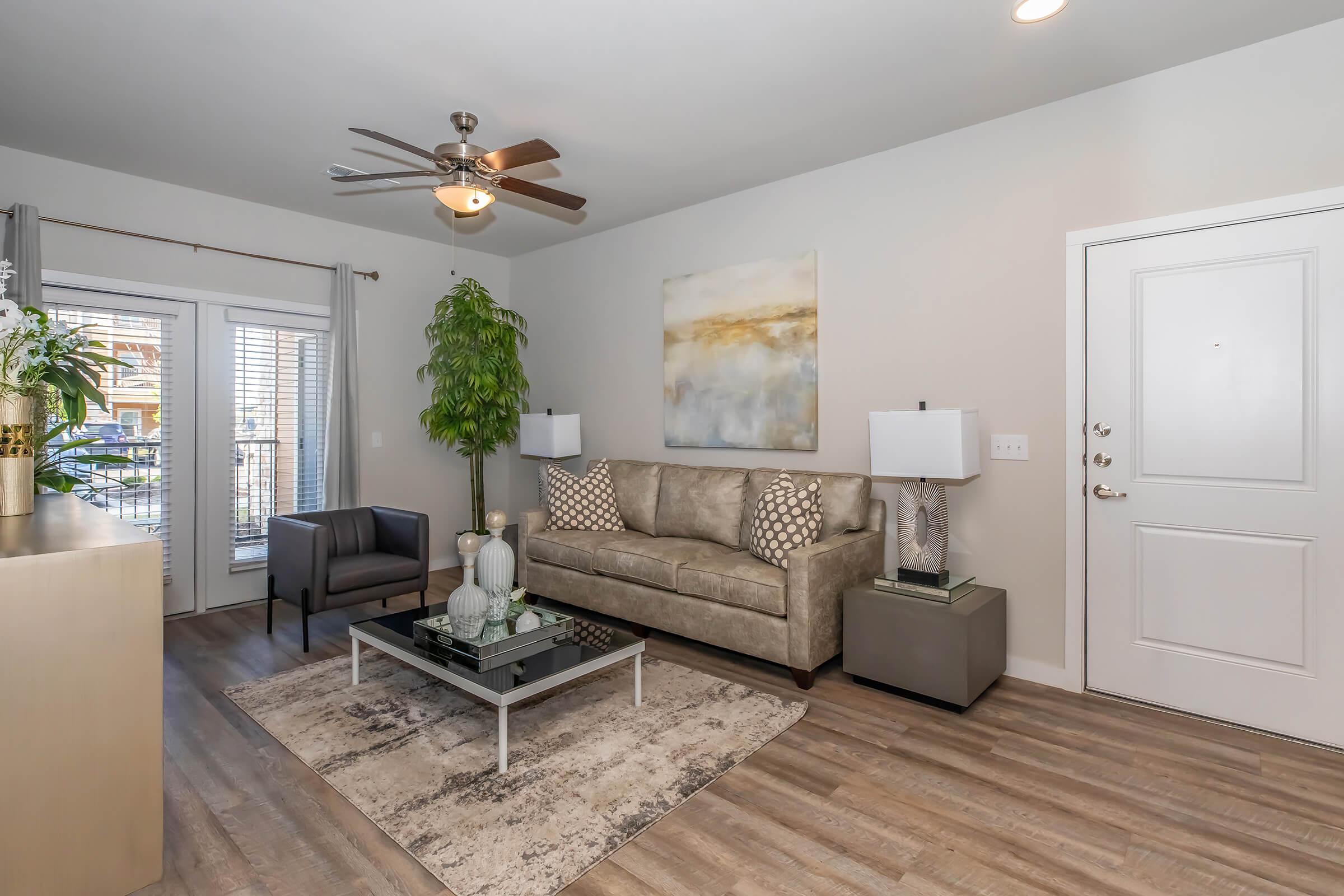 A modern living room featuring a light-colored sofa with decorative pillows, a glass coffee table, and a stylish armchair. A large abstract painting hangs on the wall, complemented by two lamps on side tables. A potted plant adds greenery, and the room has large windows with natural light.