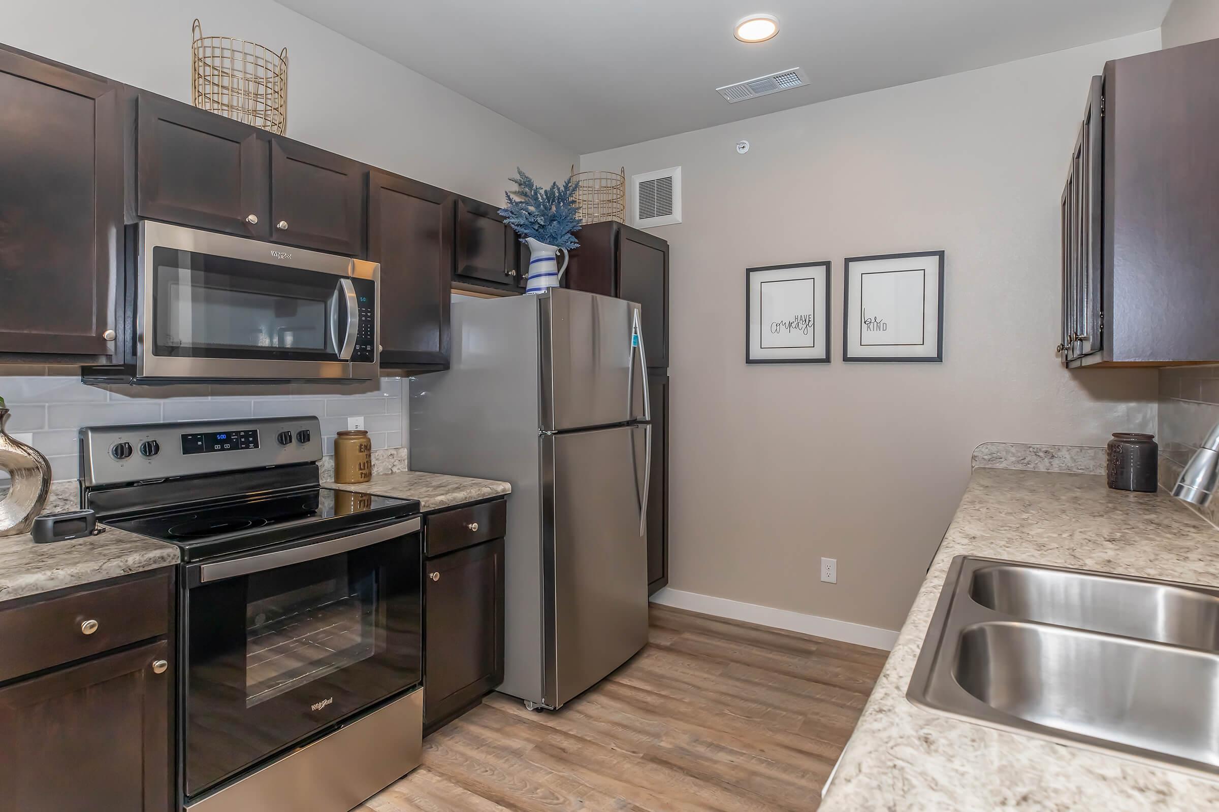 Modern kitchen featuring dark wood cabinets, stainless steel appliances including a microwave and refrigerator, and a granite countertop. There's a double sink and decorative elements like a plant and framed wall art. The flooring is wood-like, adding warmth to the space.