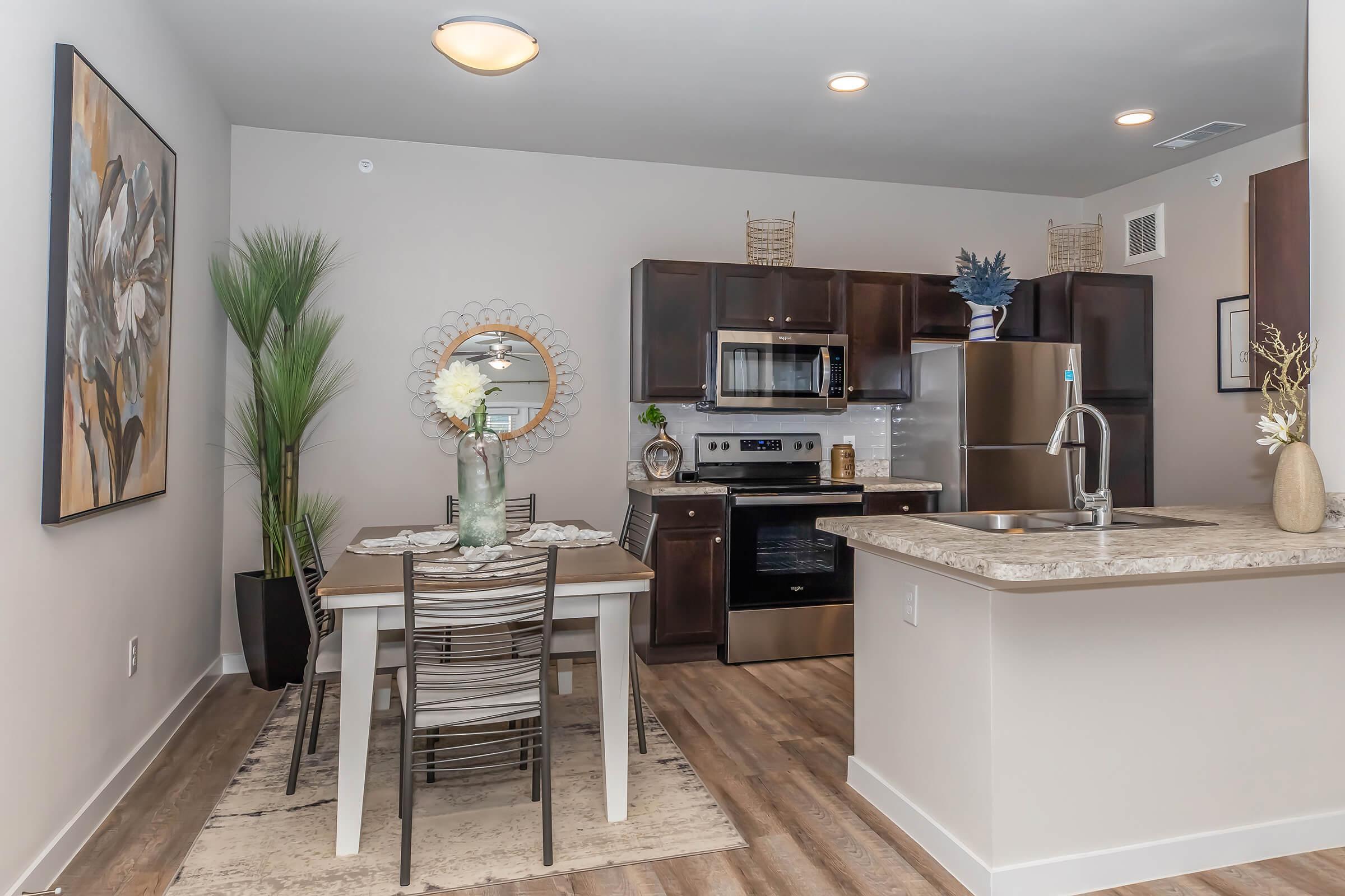Modern kitchen and dining area featuring a wooden table with chairs, stainless steel appliances, and a decorative vase. The walls are painted in neutral tones, complemented by a large artwork piece and potted plants. Natural light illuminates the space, creating a welcoming atmosphere.