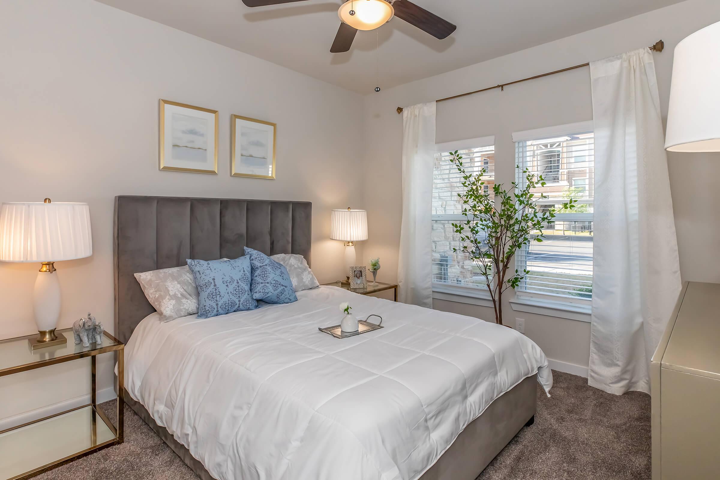 Cozy bedroom featuring a gray upholstered bed with white bedding and decorative blue pillows. Two bedside lamps illuminate the room, complemented by a small table with a decorative plant. Light curtains frame a bright window, enhancing the inviting atmosphere of the space.