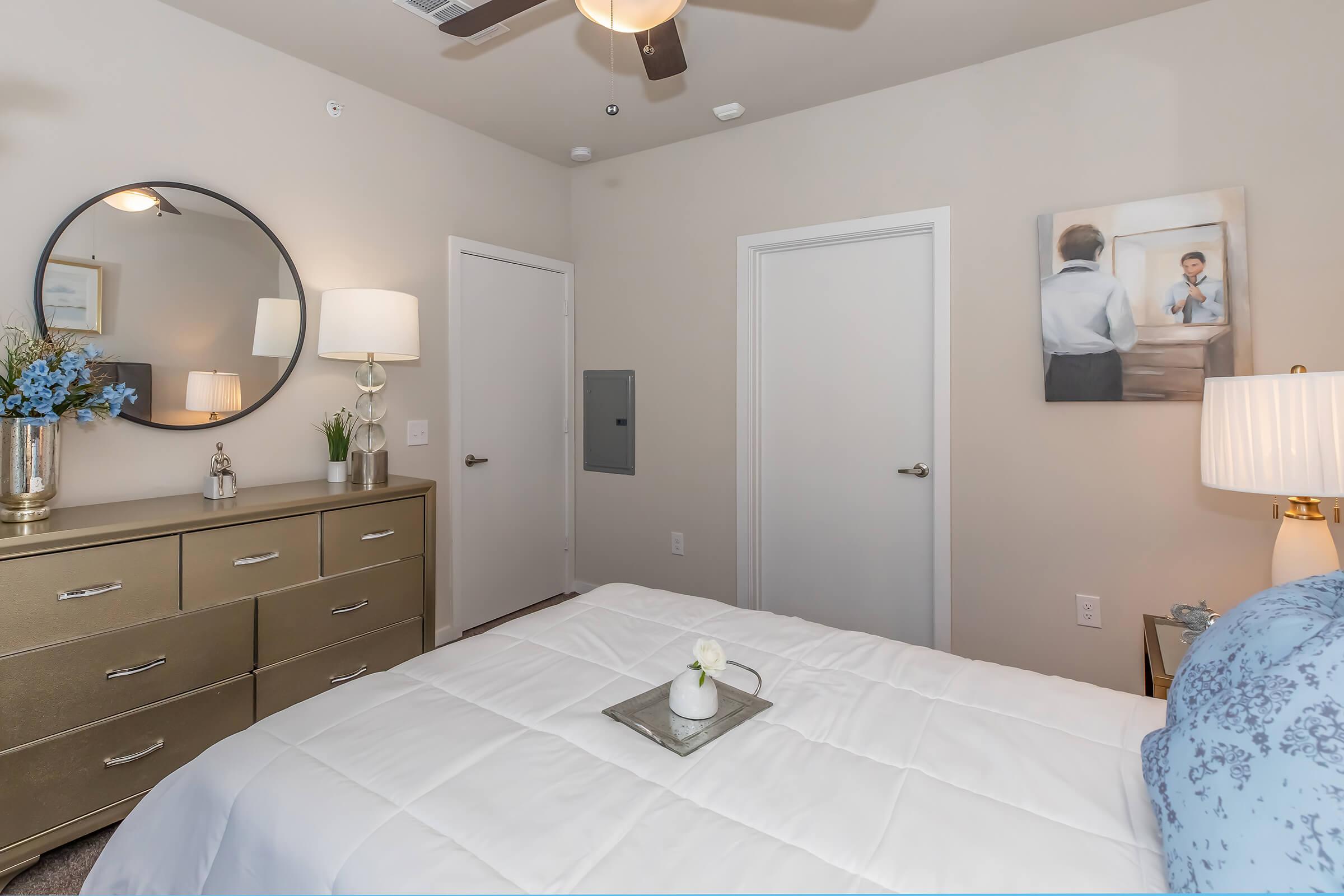 A cozy bedroom featuring a neatly made bed with a light blue blanket, a decorative tray on top, a stylish dresser with a round mirror and a lamp. There is a white door and a wall painting depicting a man in a professional setting. Soft lighting creates a warm ambiance.
