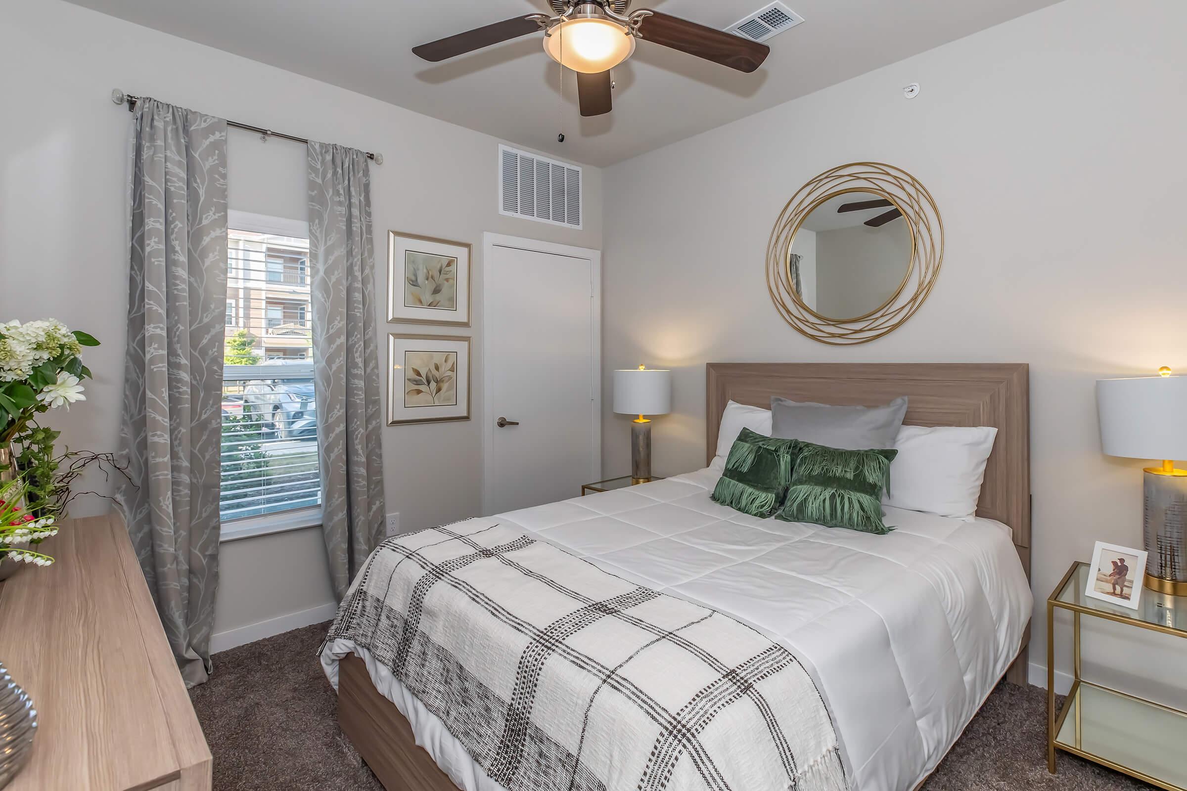 Cozy bedroom featuring a queen-sized bed with white bedding and green accent pillows. There's a wooden headboard, two stylish bedside lamps, and framed artwork on the wall. A mirror with a circular design hangs above the bed, and light gray curtains complement the interior. A dresser with decorative flowers completes the space.