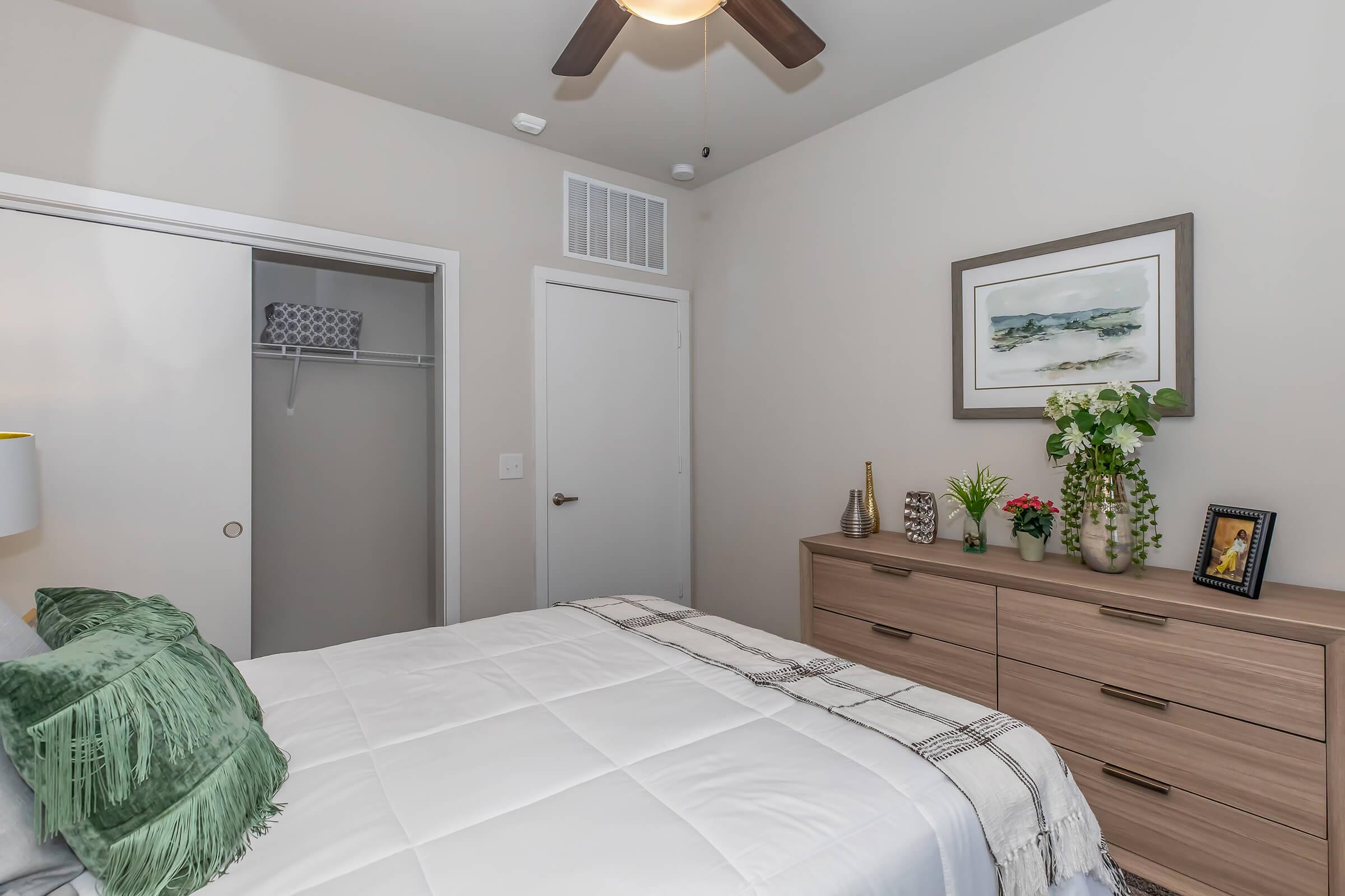 A cozy bedroom featuring a neatly made bed with green decorative pillows, a wooden dresser adorned with plants and a photo frame, and a closet with sliding doors. The walls are a soft beige, and a ceiling fan adds a functional touch. Natural light enhances the inviting atmosphere.