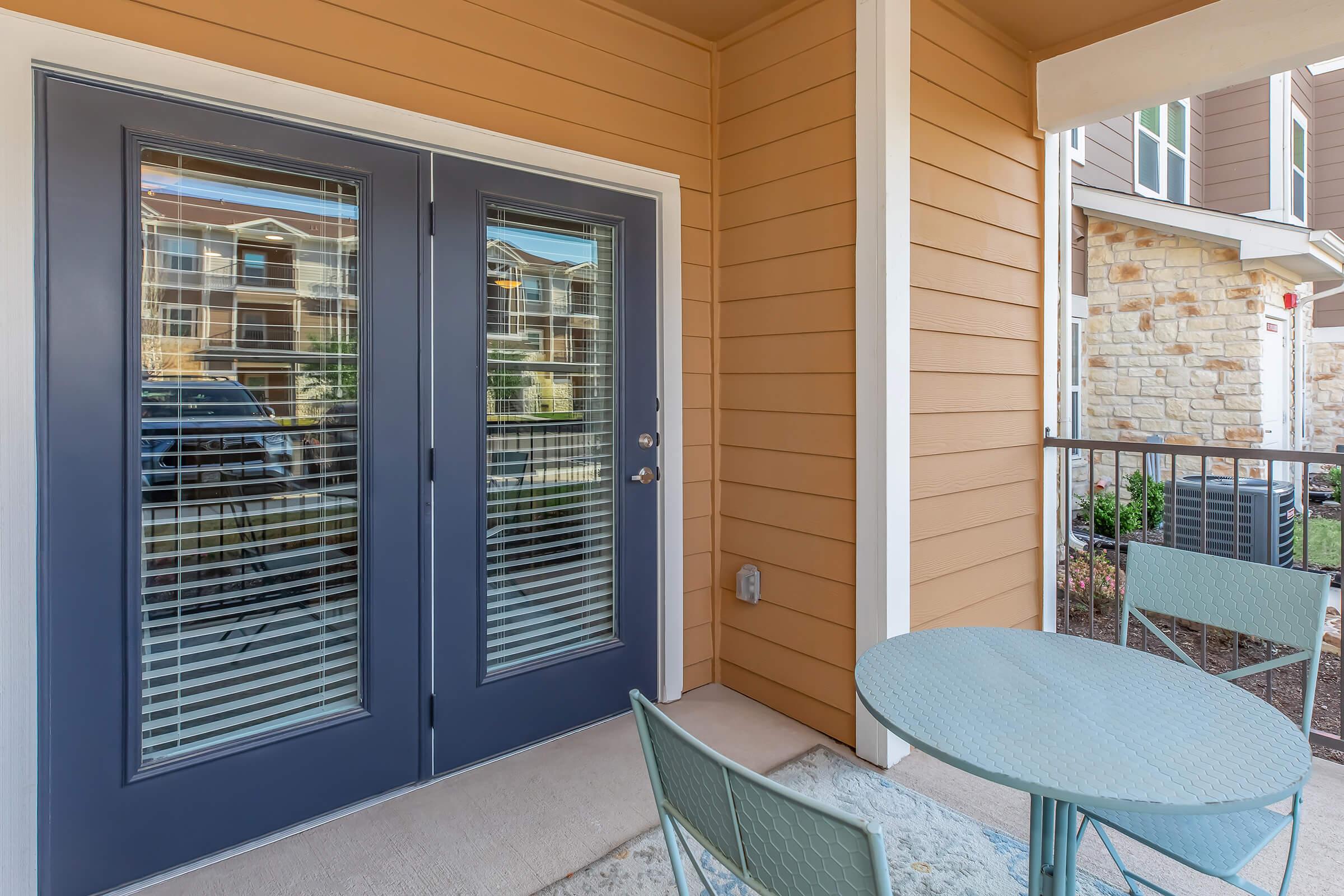 A cozy porch area featuring a set of double doors with blinds, a round table, and two chairs. The walls are painted in warm tones, and the floor is covered with a mat. In the background, there are hints of a landscaped area and nearby buildings.
