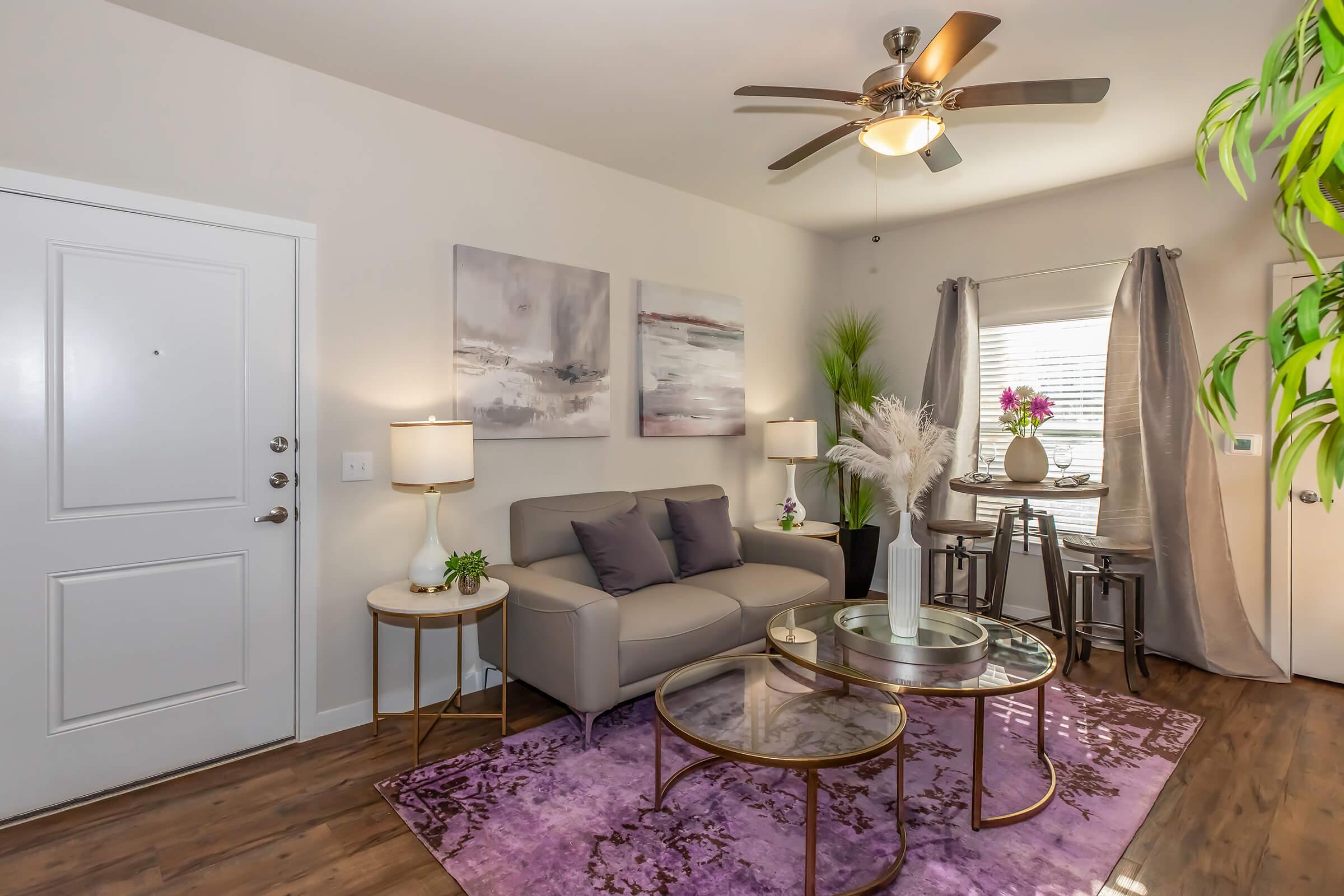 A modern living room featuring a grey sofa with cushions, two round glass coffee tables, and a vibrant purple area rug. There are two stylish lamps placed on side tables, and a small dining table with chairs in the corner. The decor includes artwork and plants, creating a cozy and inviting atmosphere.