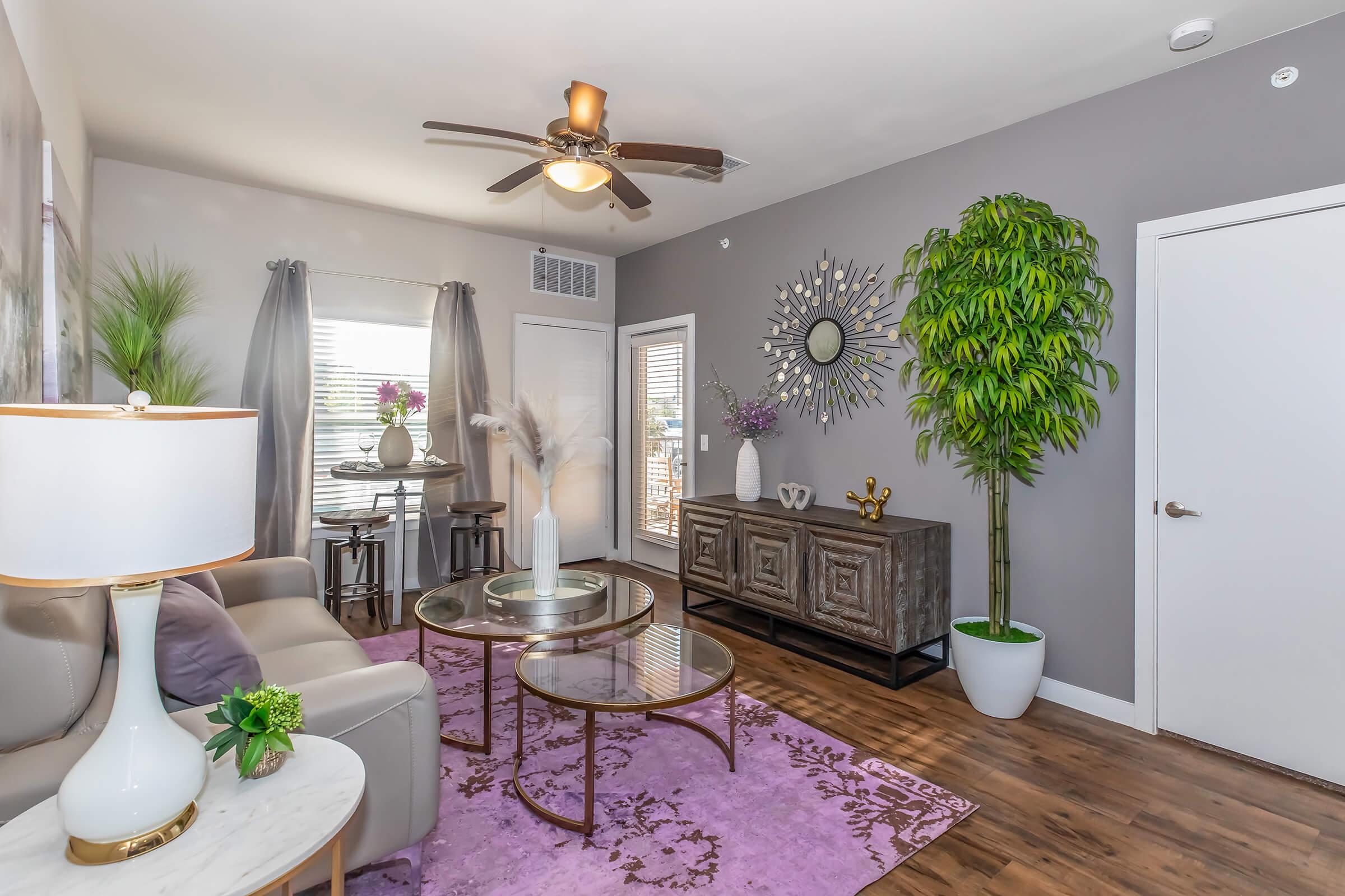 A modern living room featuring a light-colored sofa, a round dining table with chairs, and decorative plants. The walls are painted gray, adorned with a large mirror and wall art. A stylish cabinet holds decorative items, while a purple rug adds a pop of color to the wooden floor. Natural light enters through bright windows.