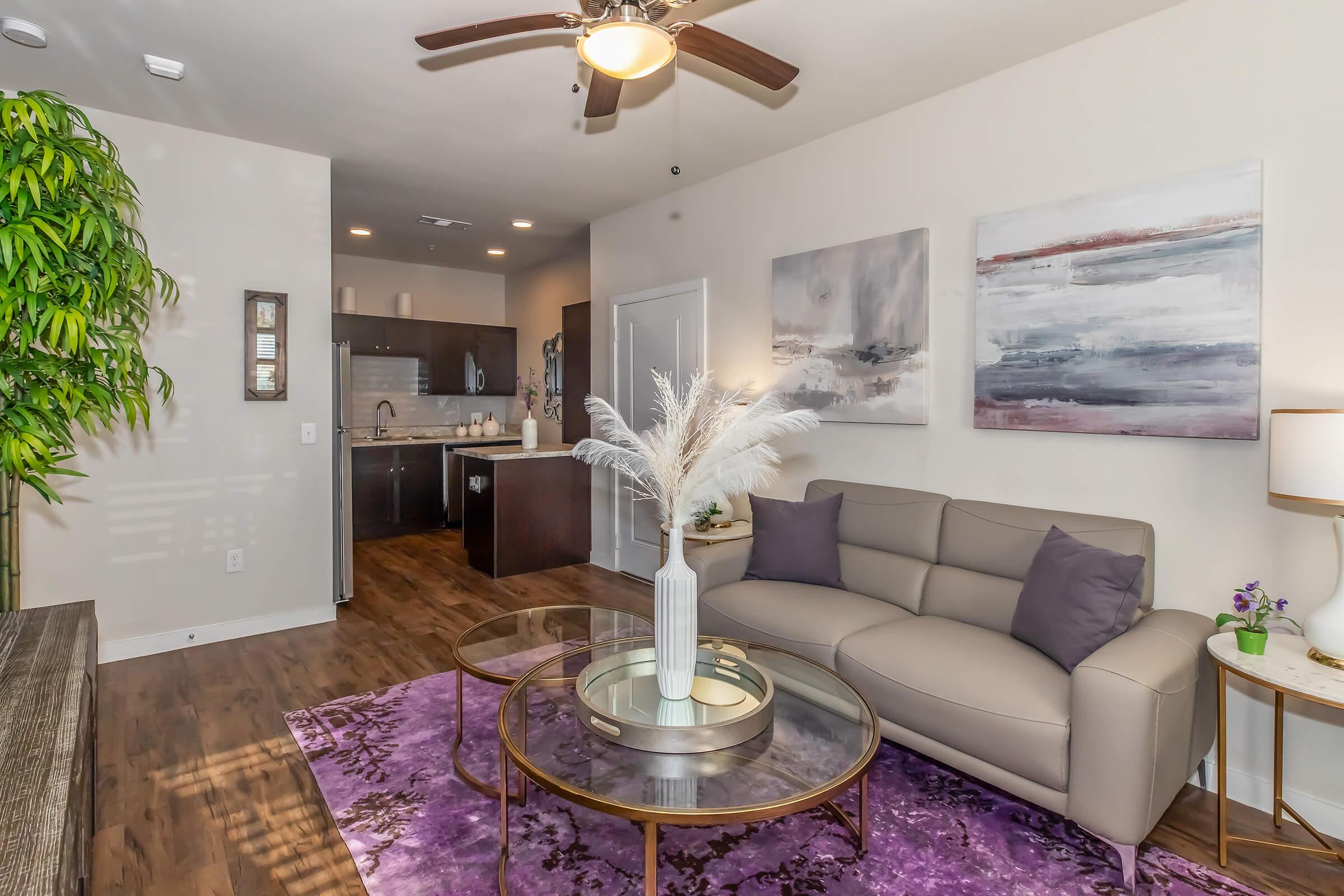 A modern living room featuring a light gray sofa with purple cushions, a round glass coffee table, and a decorative vase with white feathers. The space has a purple area rug and two abstract paintings on the wall. A kitchen area is visible in the background, and a tall green plant adds a touch of nature.
