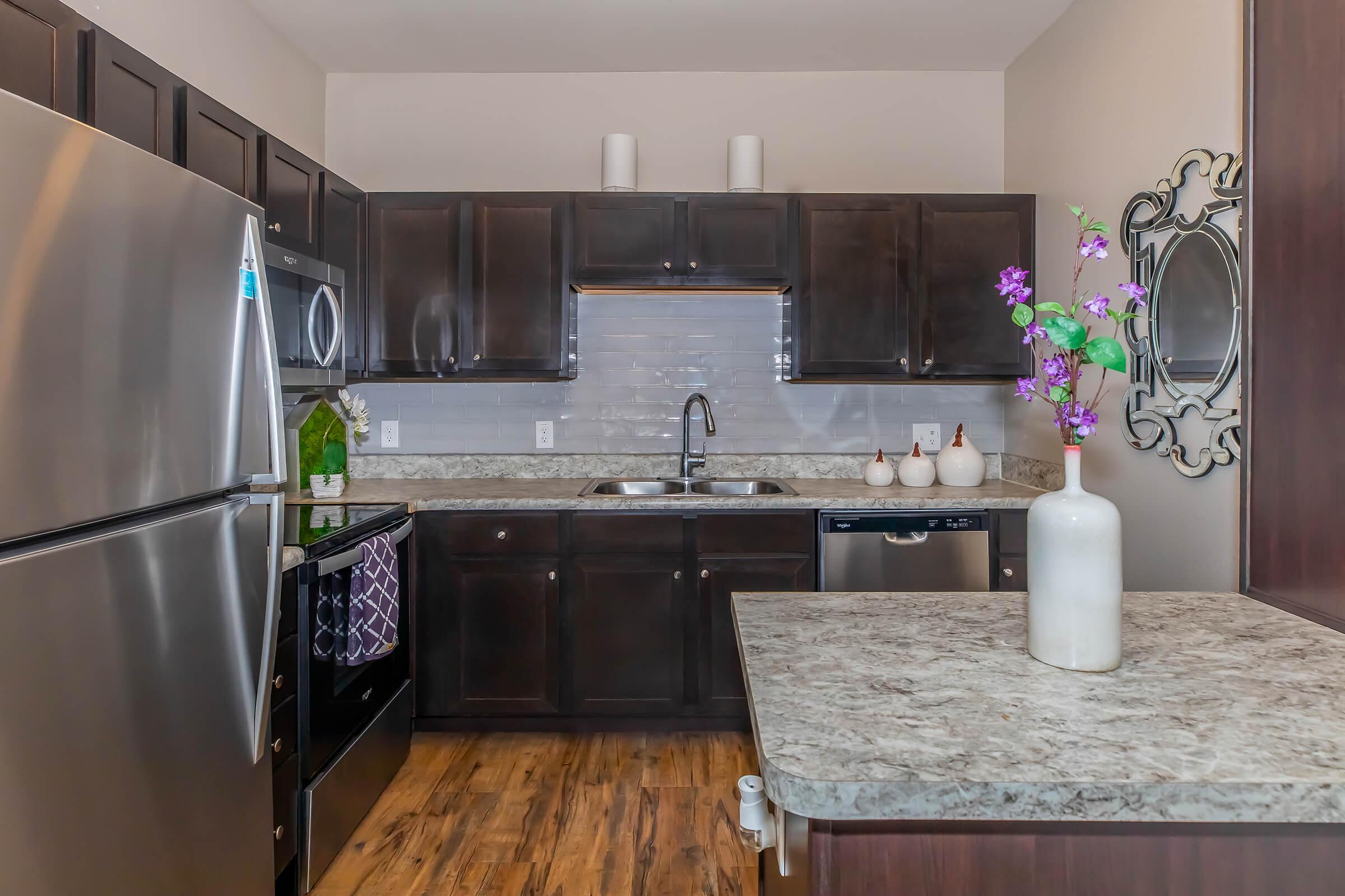 A modern kitchen featuring dark wooden cabinetry, a stainless steel refrigerator, oven, and dishwasher. The countertop is speckled, and there's a vase with purple flowers on the island. The backsplash is tiled in light colors, and decorative items are arranged on the shelves.