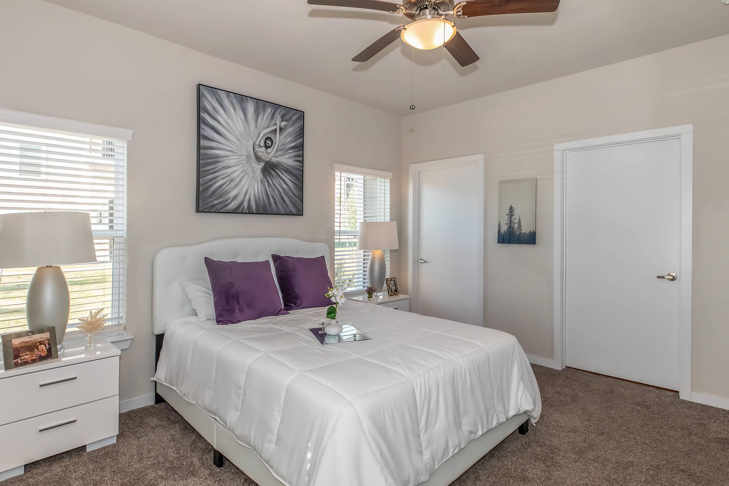 A cozy bedroom featuring a queen-sized bed with white bedding and purple accent pillows. There are two bedside lamps on white nightstands, a wall art piece depicting an eye, and a large window with blinds. The room has light-colored walls and a soft carpet, creating a warm atmosphere.