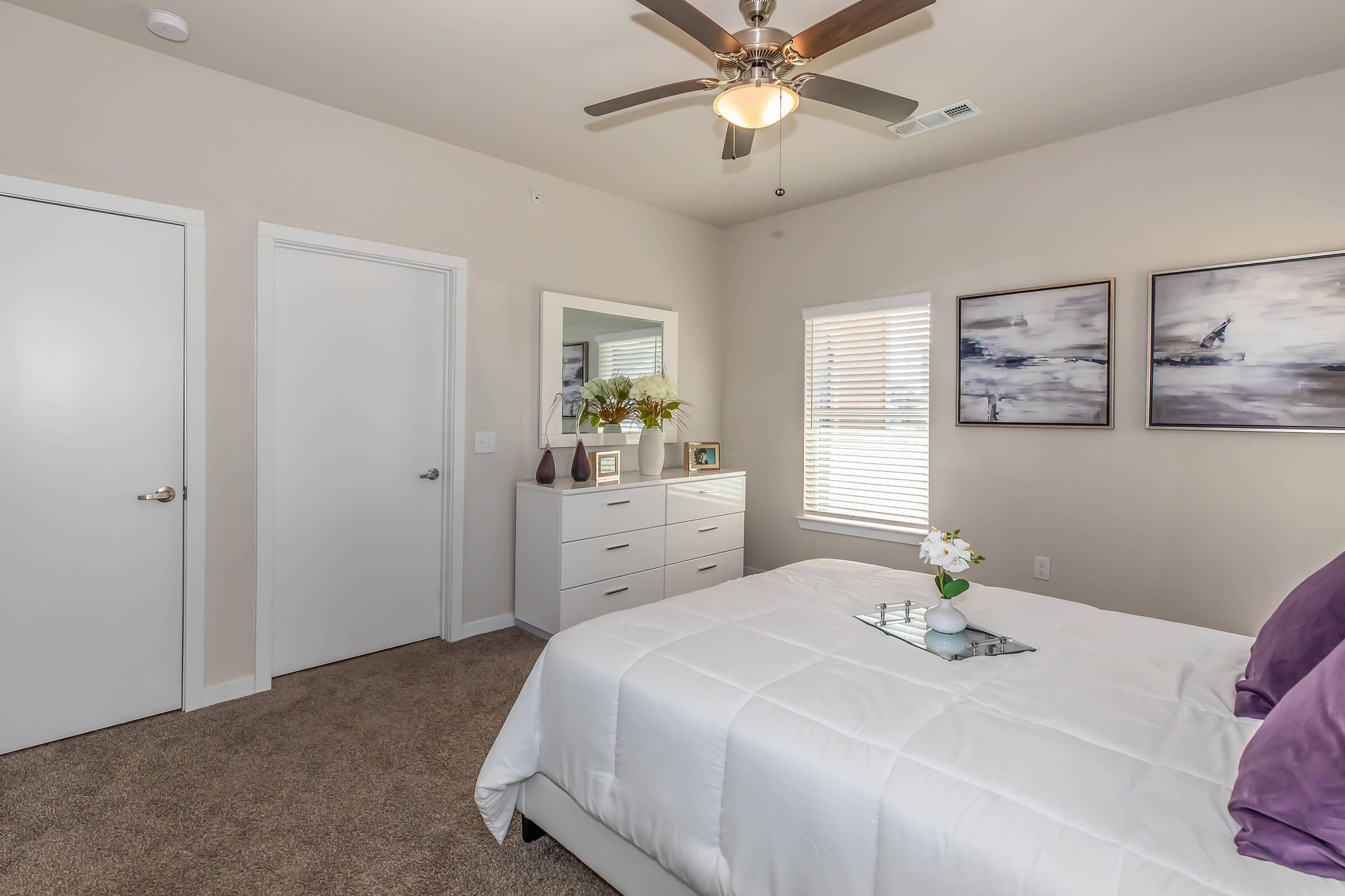 A well-decorated bedroom featuring a bed with white bedding and purple throw pillows. There is a nightstand with decor on top, a full-length mirror, a dresser, and two framed pictures on the wall. A window with blinds allows natural light in. The room has light-colored walls and carpeted flooring.