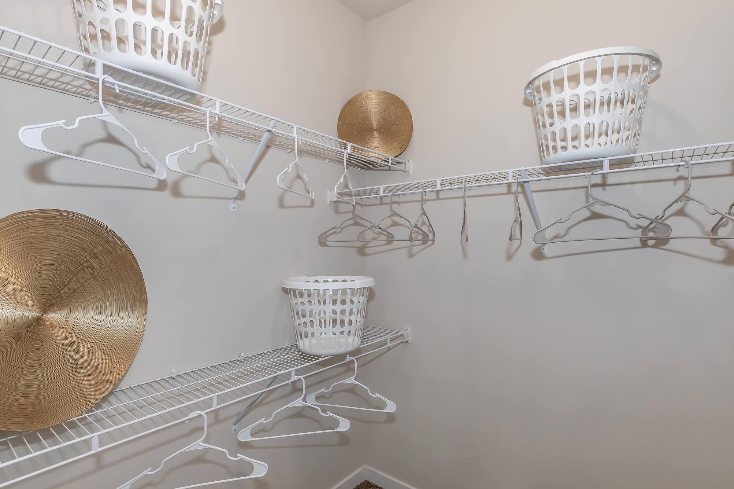 A laundry room space featuring white wire shelving with several hanging clothes hangers. Two round, gold decorative plates are placed on the shelves, along with two white laundry baskets. The walls are a light neutral color, and the floor is carpeted. The area appears well-organized and tidy.