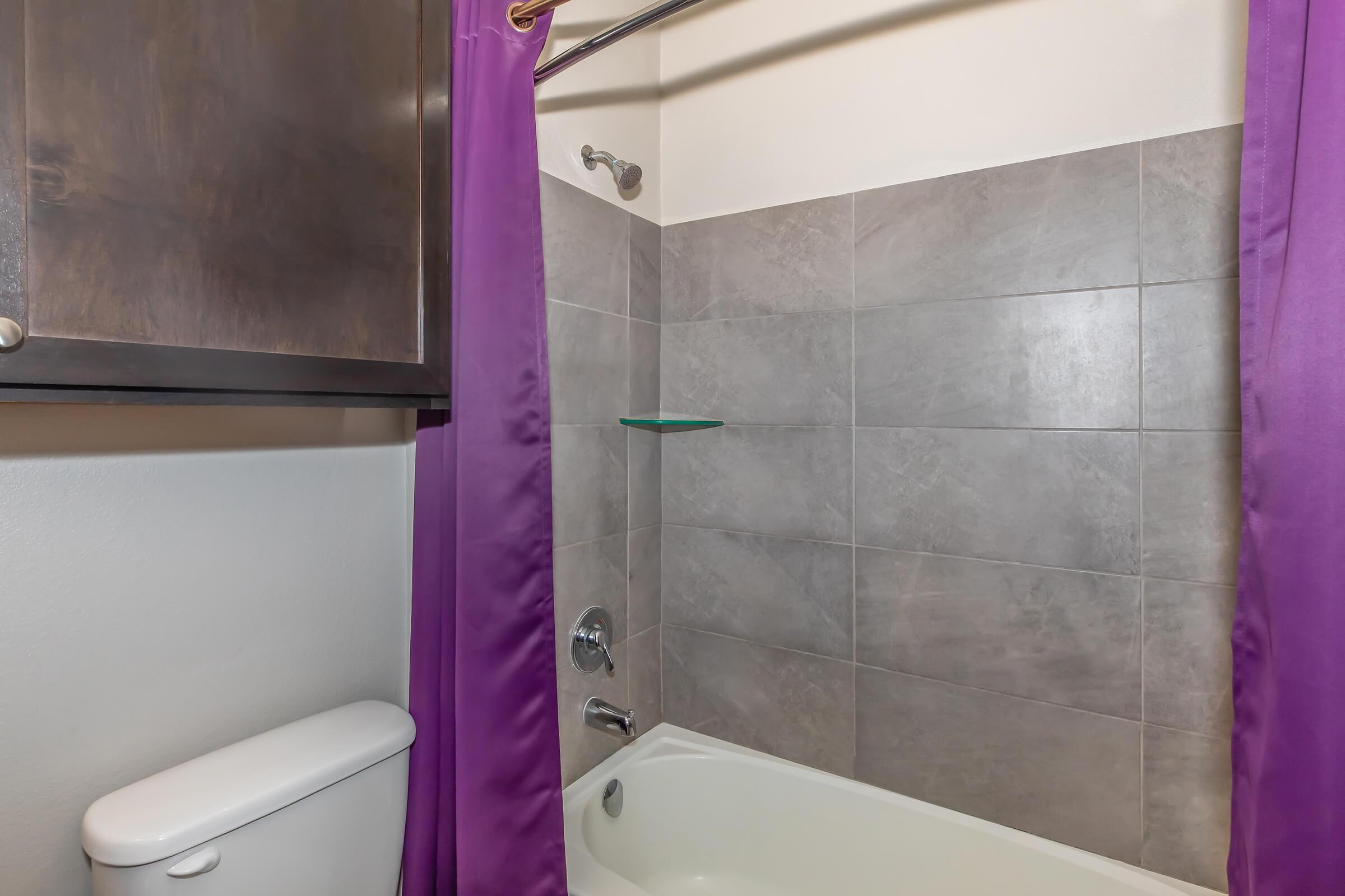 A bathroom featuring a bathtub with a shower, a purple shower curtain, and gray tiled walls. A white toilet is visible in the foreground, along with a glass shelf on the wall. The overall design is modern and clean, with a neutral color palette.