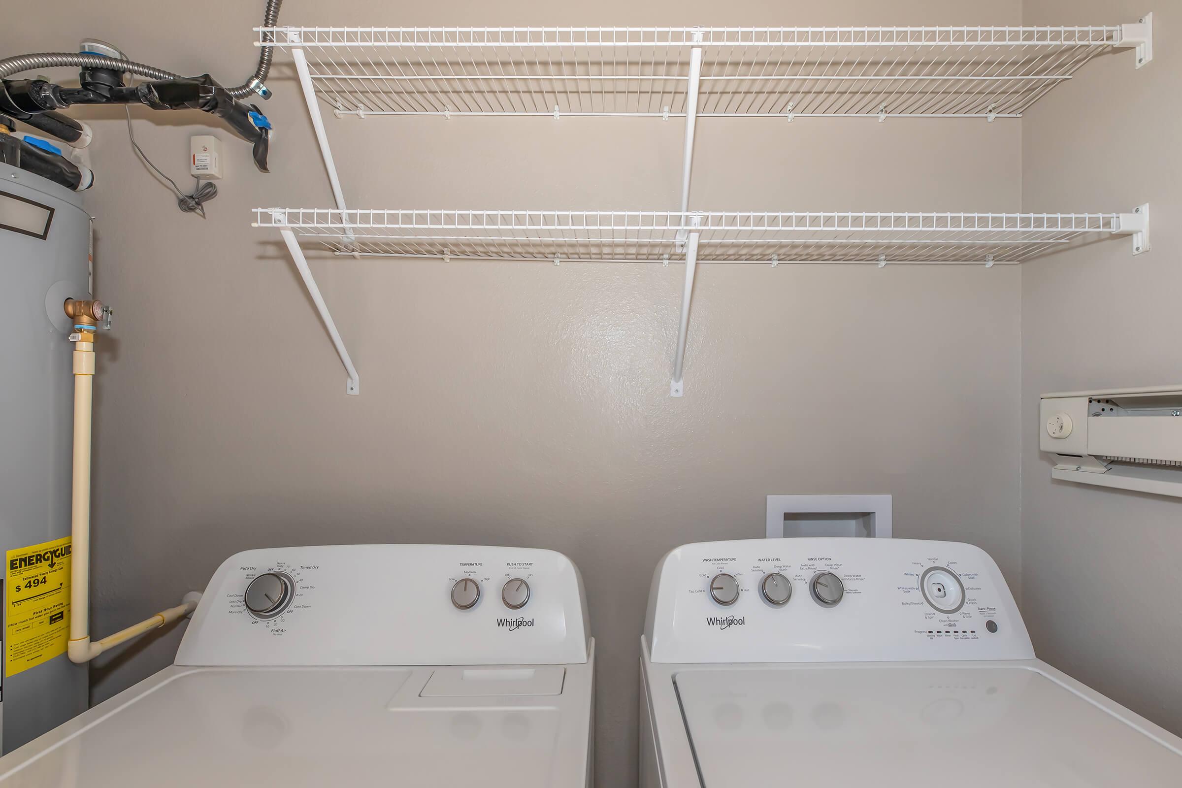 A laundry room featuring a white washing machine and dryer side by side, with control panels visible. Above them, there are wire shelving units mounted on the wall for storage. A water heater is located on the left side of the image, partially visible. The walls are painted in a light neutral color.