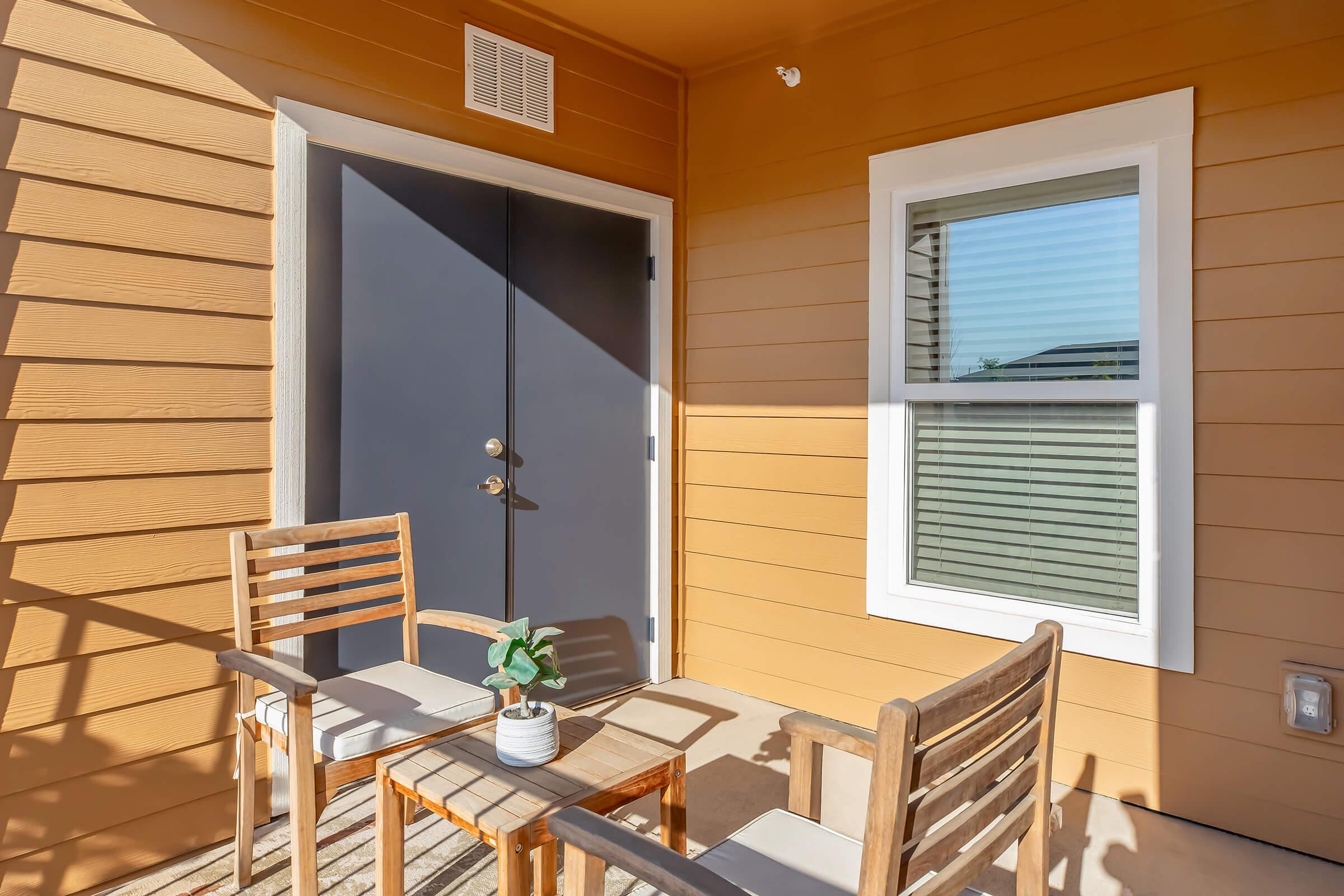 A small outdoor patio featuring two wooden chairs and a table with a potted plant. The patio has a beige wooden wall and a dark gray door leading inside. There's a window on one side with horizontal blinds, allowing sunlight to filter in.