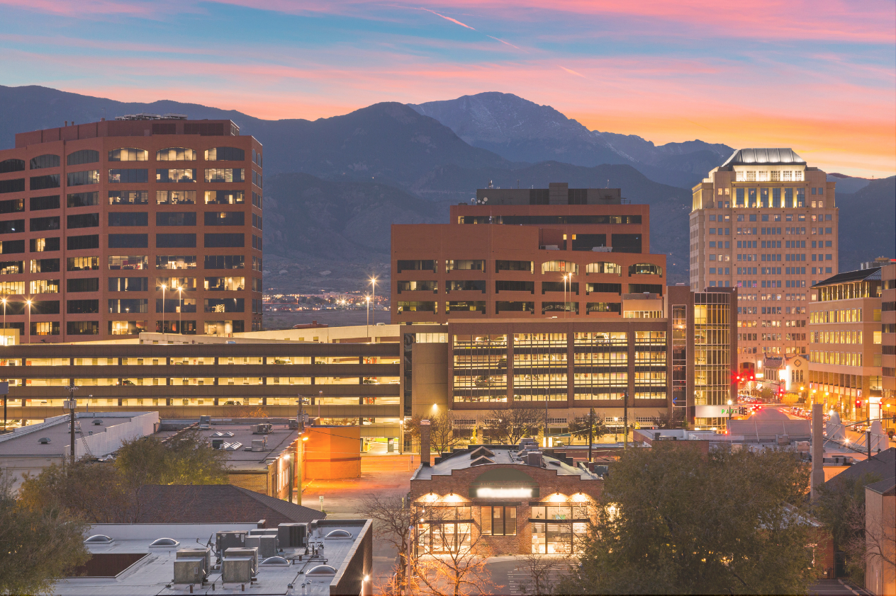 A vibrant sunset over a city skyline, featuring modern buildings and a mountain backdrop. The sky is painted in shades of pink and orange, while lights begin to twinkle in the urban landscape below. The image captures the transition from day to night in a bustling downtown area.