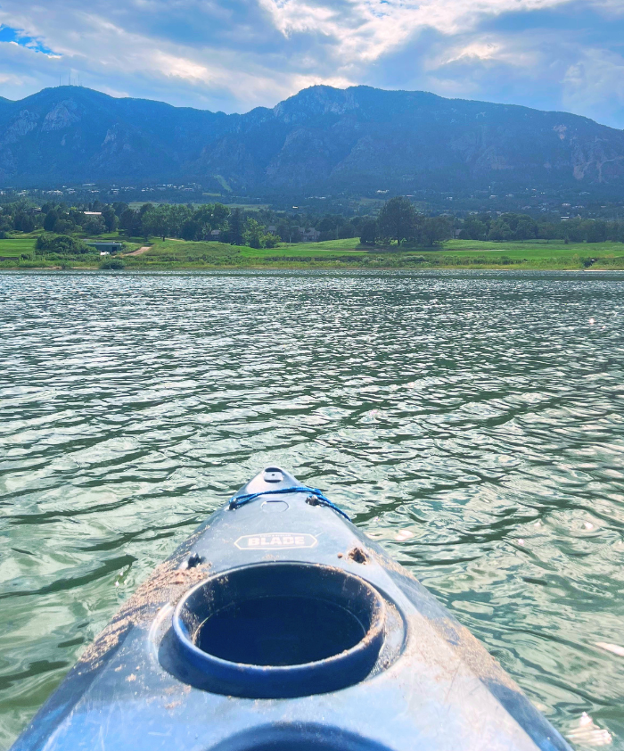 View from a kayak on calm water, with mountains in the background under a partly cloudy sky. The kayak's cockpit is visible in the foreground, and lush green fields are seen along the shoreline. The scene conveys a peaceful outdoor experience on a lake.