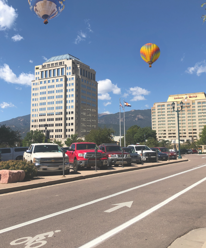 A city street scene featuring a bike lane lined with parked cars. In the background, there are tall buildings and a clear blue sky. Hot air balloons are floating overhead, adding a colorful touch to the urban landscape. The scene conveys a vibrant day in a bustling city.