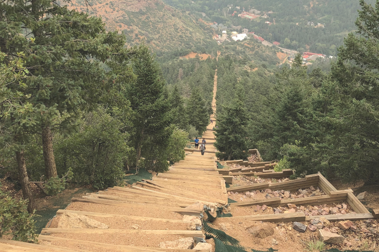 A steep wooden staircase winding down through dense pine trees, leading to a valley below. Two hikers can be seen making their way down the stairs, with lush green surroundings and a distant view of a small town nestled in the landscape. The path appears well-maintained and invites outdoor exploration.