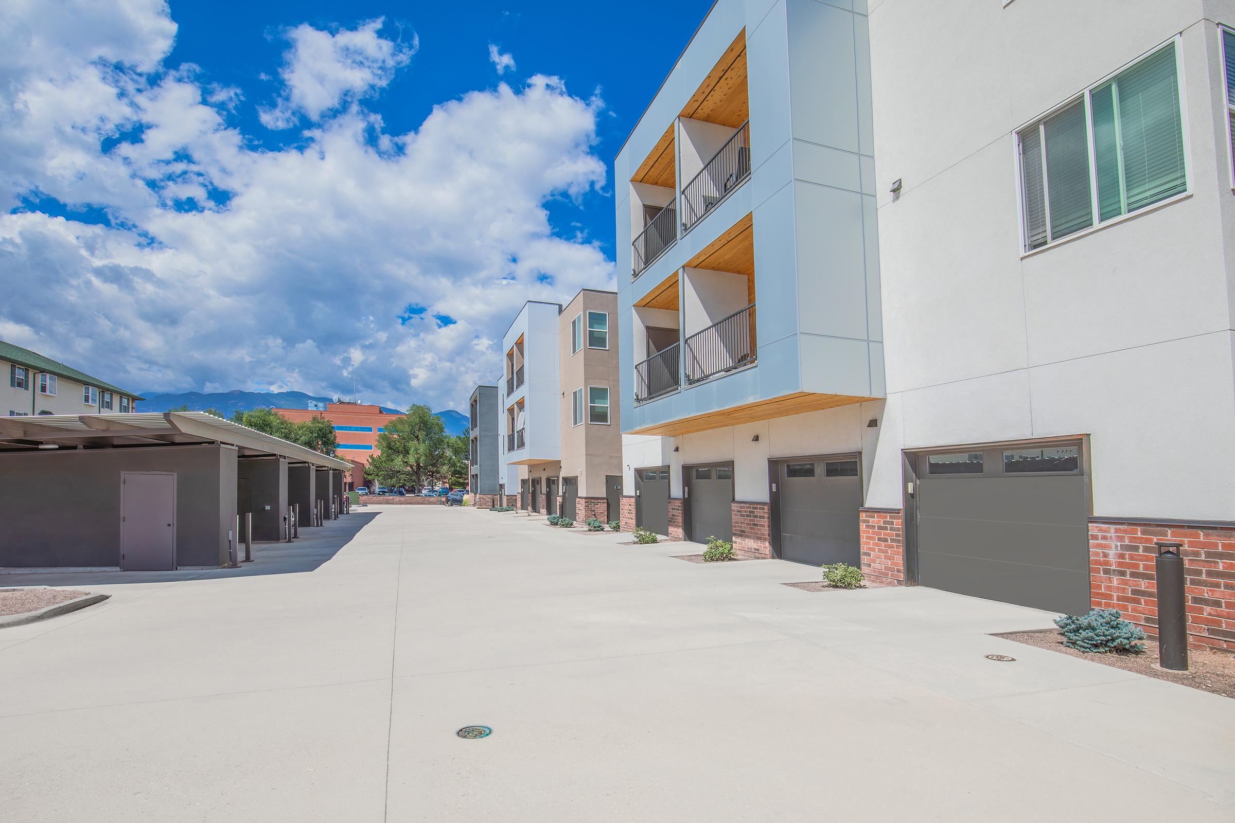 Modern residential buildings with balconies and garages along a clean, paved pathway. The sky is bright with scattered clouds, and there are trees in the background, indicating a sunny day. Several parked vehicles and landscaped areas can also be seen, contributing to an inviting neighborhood atmosphere.