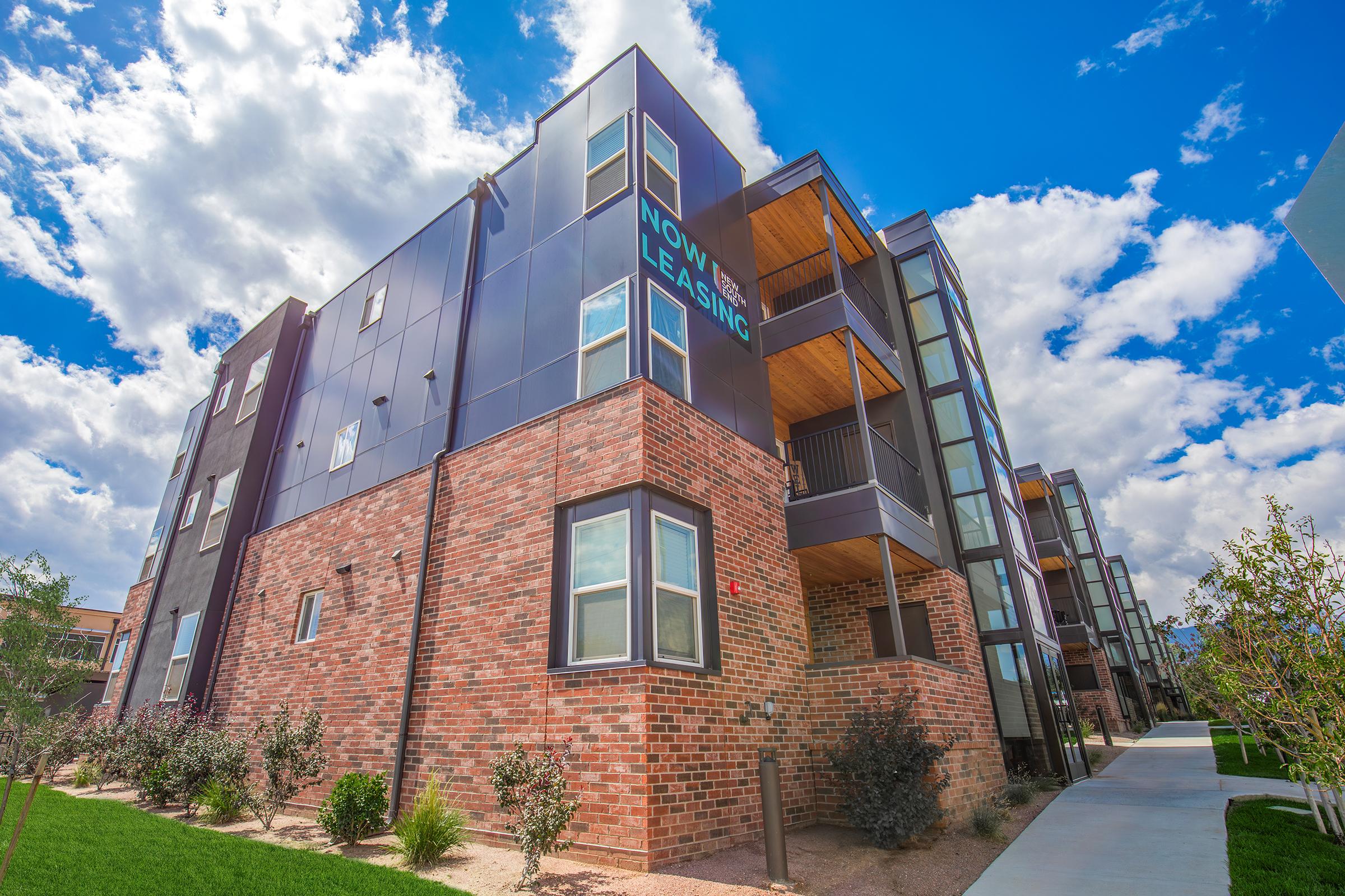 Modern apartment building with a mix of brick and metal exteriors. The structure features multiple levels, large windows, and a "Now Leasing" sign prominently displayed. The background includes a bright blue sky with white clouds, and the foreground has landscaped greenery and a walkway.