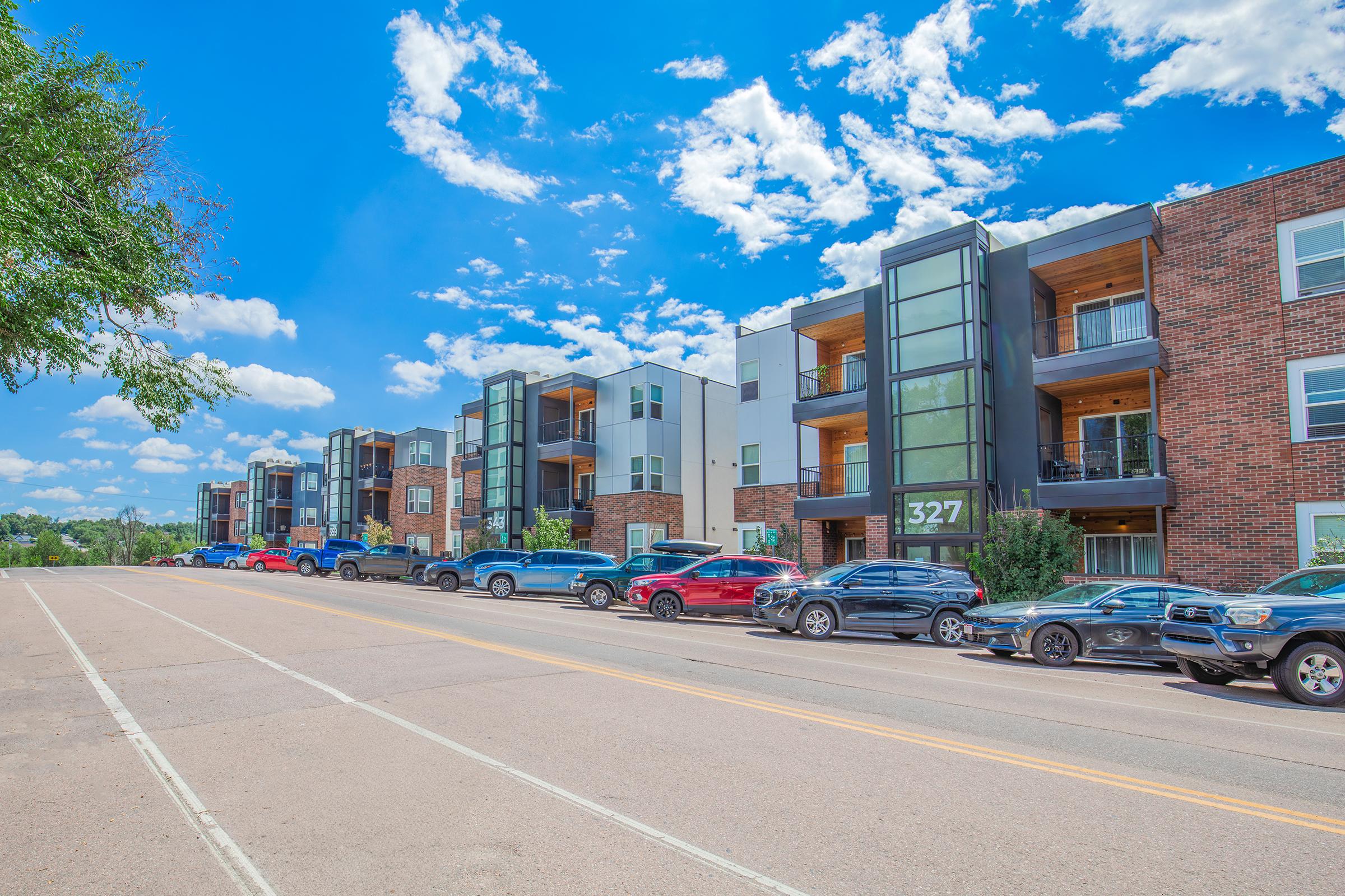 Modern apartment buildings line a sunny street, with a clear blue sky overhead and fluffy white clouds. Several parked cars are visible along the road. The front of one building features large windows and a prominent address number 327. Greenery adds a touch of nature to the urban scene.