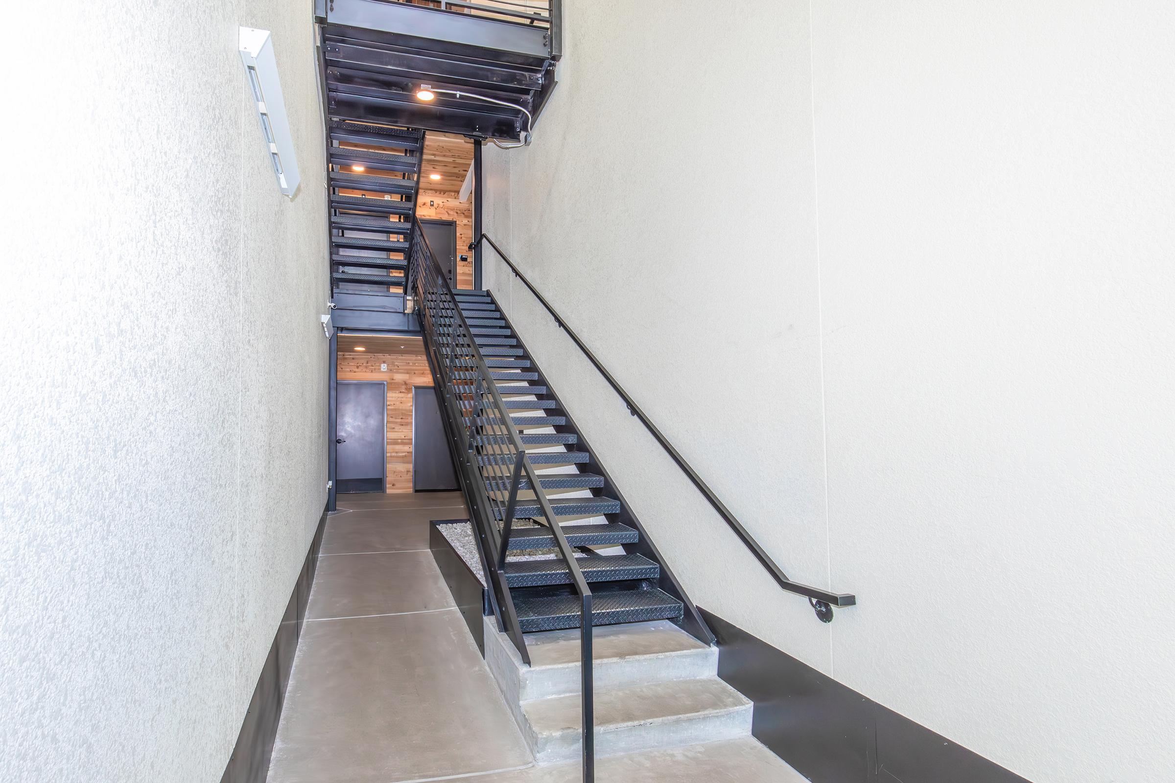 Interior view of a modern staircase in an industrial-style building. Black metal railing along the stairs, which are surrounded by light gray walls. Natural light from overhead fixtures illuminates the space, highlighting the combination of metal and wood elements.