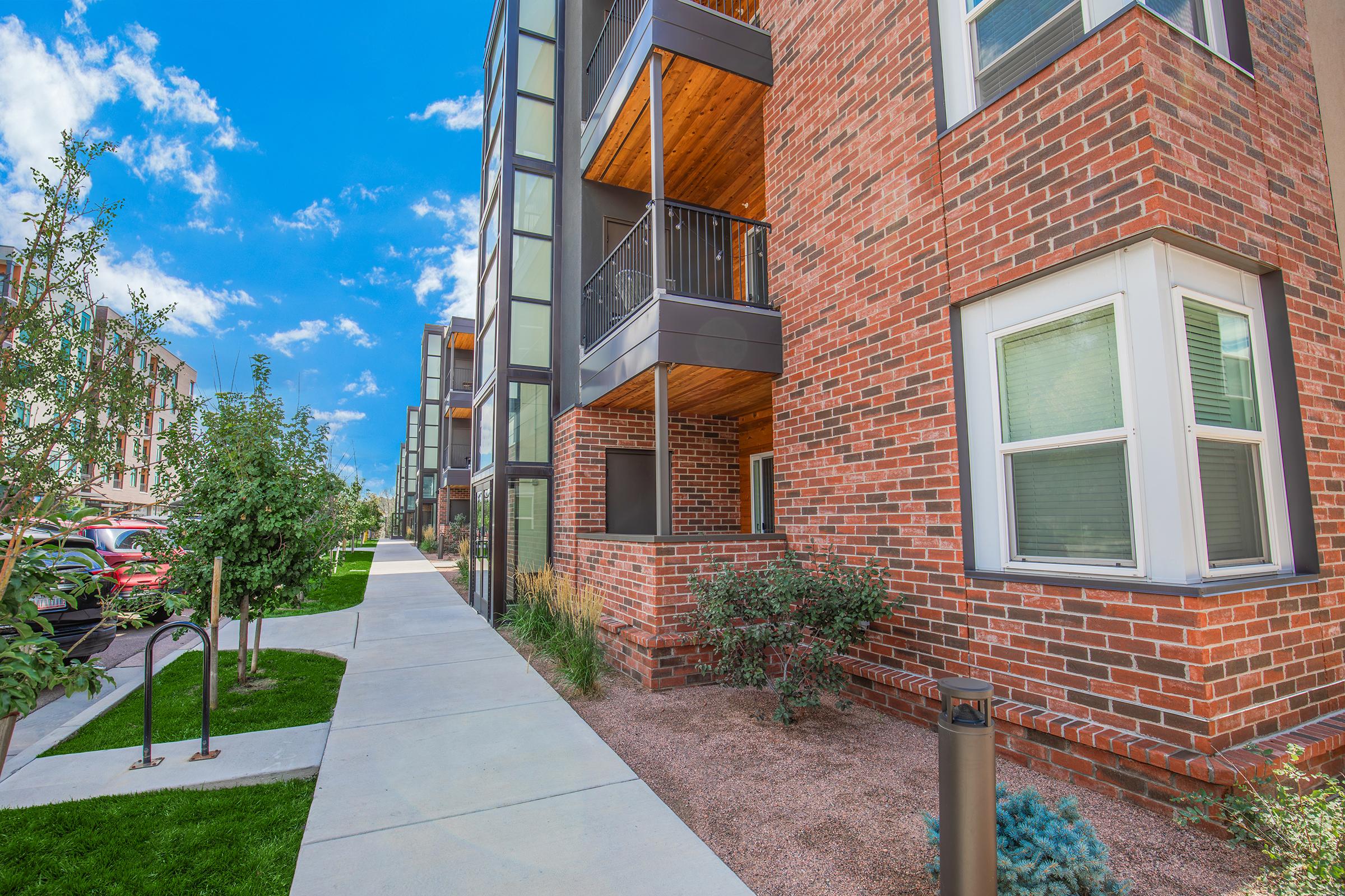 A modern apartment building made of brick with large windows and balconies, situated alongside a paved sidewalk. Lush greenery, including small trees and shrubs, adorns the area, while blue skies with scattered clouds create a bright, inviting atmosphere.