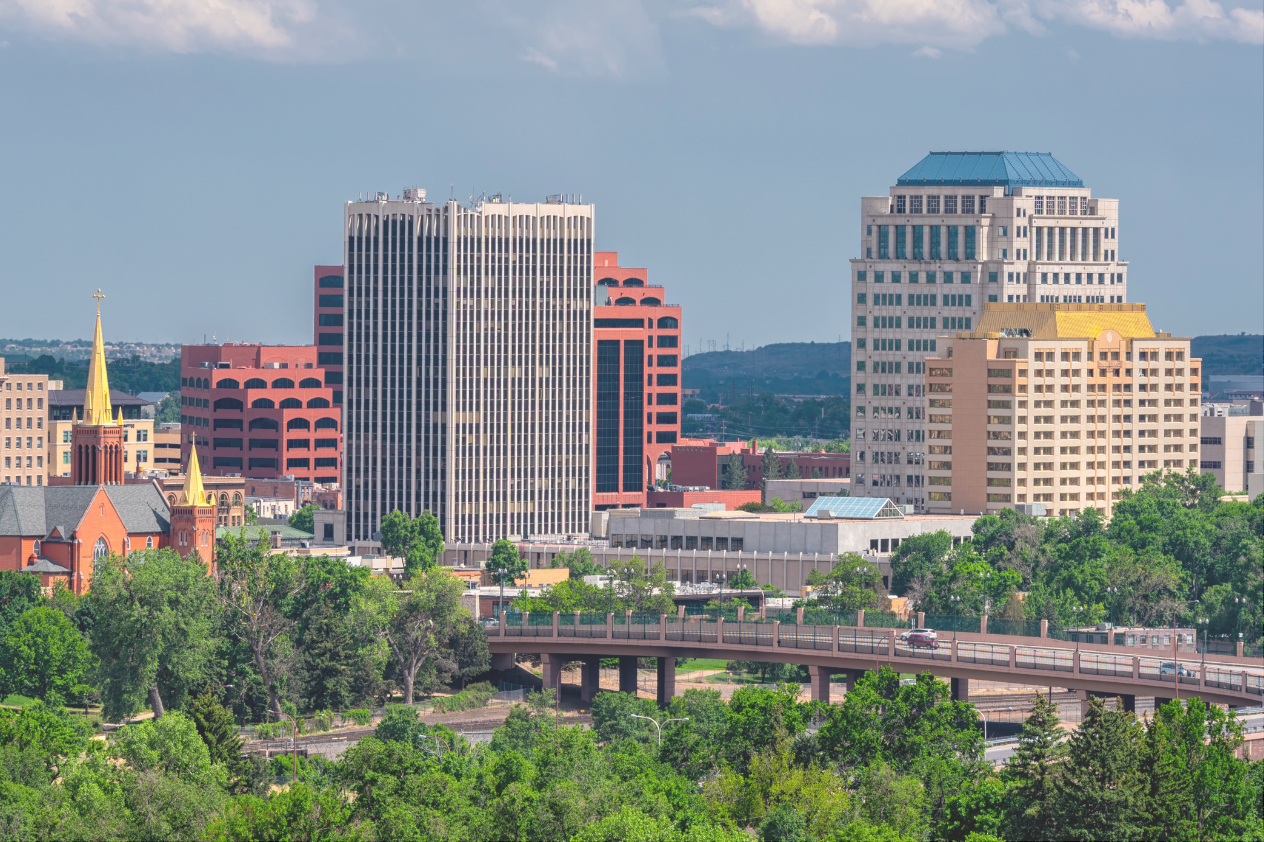 Skyline view of a city featuring a mix of modern and historical buildings, including tall office towers and a church with a steeple. Lush green trees line the foreground, and a highway runs beneath the buildings, under a partly cloudy sky. The city exhibits a blend of architectural styles.