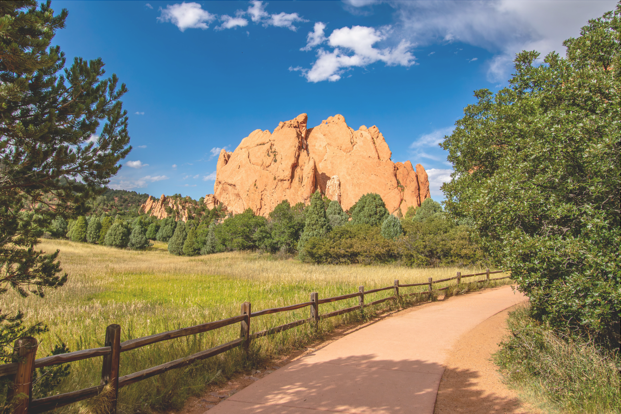 A scenic view of a rocky formation surrounded by greenery, with a clear blue sky and scattered clouds. A well-maintained path lined with a wooden fence winds through a grassy area, inviting outdoor exploration.