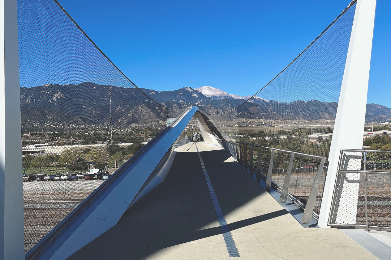 A modern pedestrian bridge with a sleek design leads towards mountains in the background, under a clear blue sky. The bridge features metal mesh sides and a smooth pathway, while the distant mountains show a hint of snow on their peaks. The scenery includes greenery and urban elements beneath the bridge.