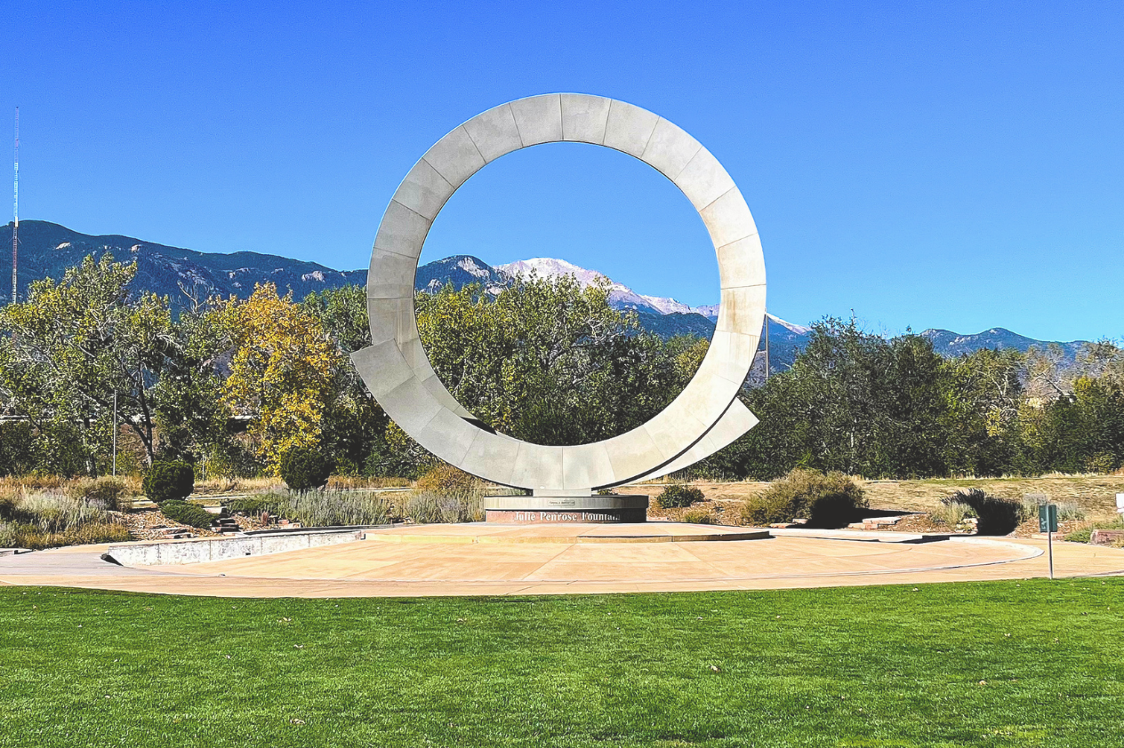 A large circular sculpture set against a clear blue sky, surrounded by green grass and trees. In the background, mountains are partially covered with snow. The sculpture features a smooth, modern design and is situated in an open park area, showcasing an artistic element that complements the natural landscape.